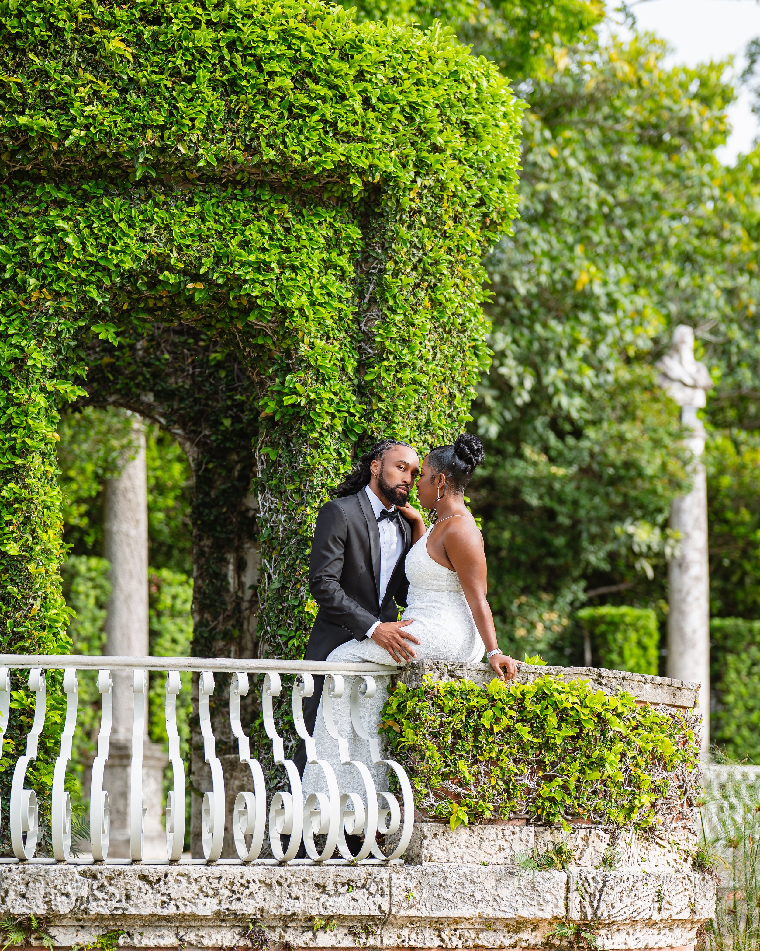 A couple dressed in wedding attire standing together on a stone and metal railing platform outdoors, with lush green trees and a statue in the background.