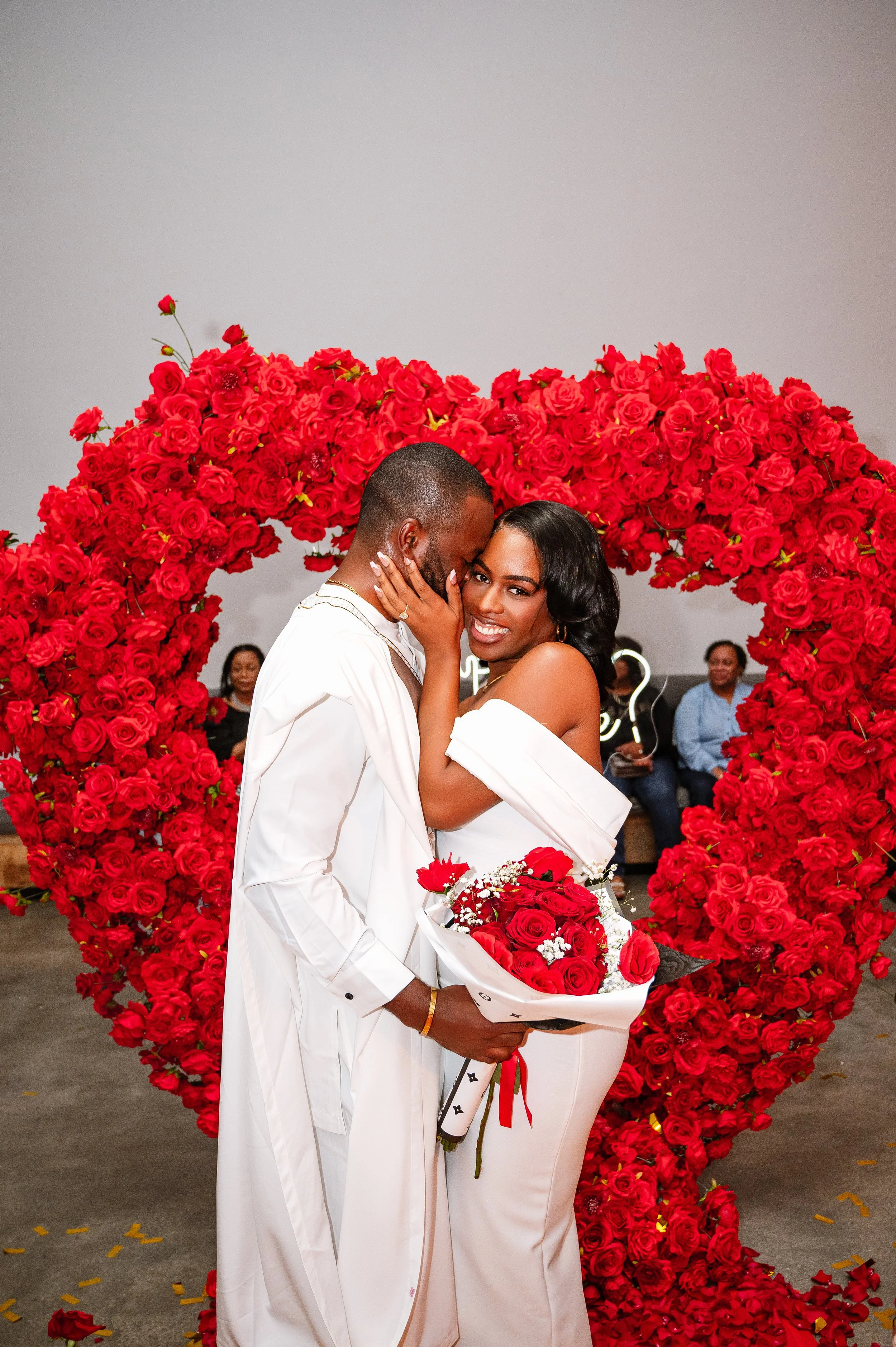 A couple, dressed in white, sharing an intimate moment in front of a large heart-shaped display made of red roses. The woman holds a bouquet of red roses wrapped in paper, and both are smiling. Several people are seated in the background observing th