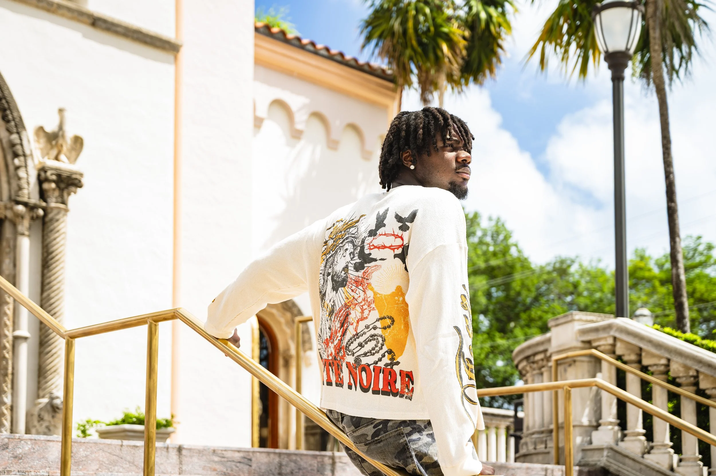 A young man with dreadlocks wearing a white sweater with colorful graphic prints walks up a staircase outside a building with a white stucco wall, arched windows, and orange trim, under a bright blue sky with some clouds, surrounded by palm trees.