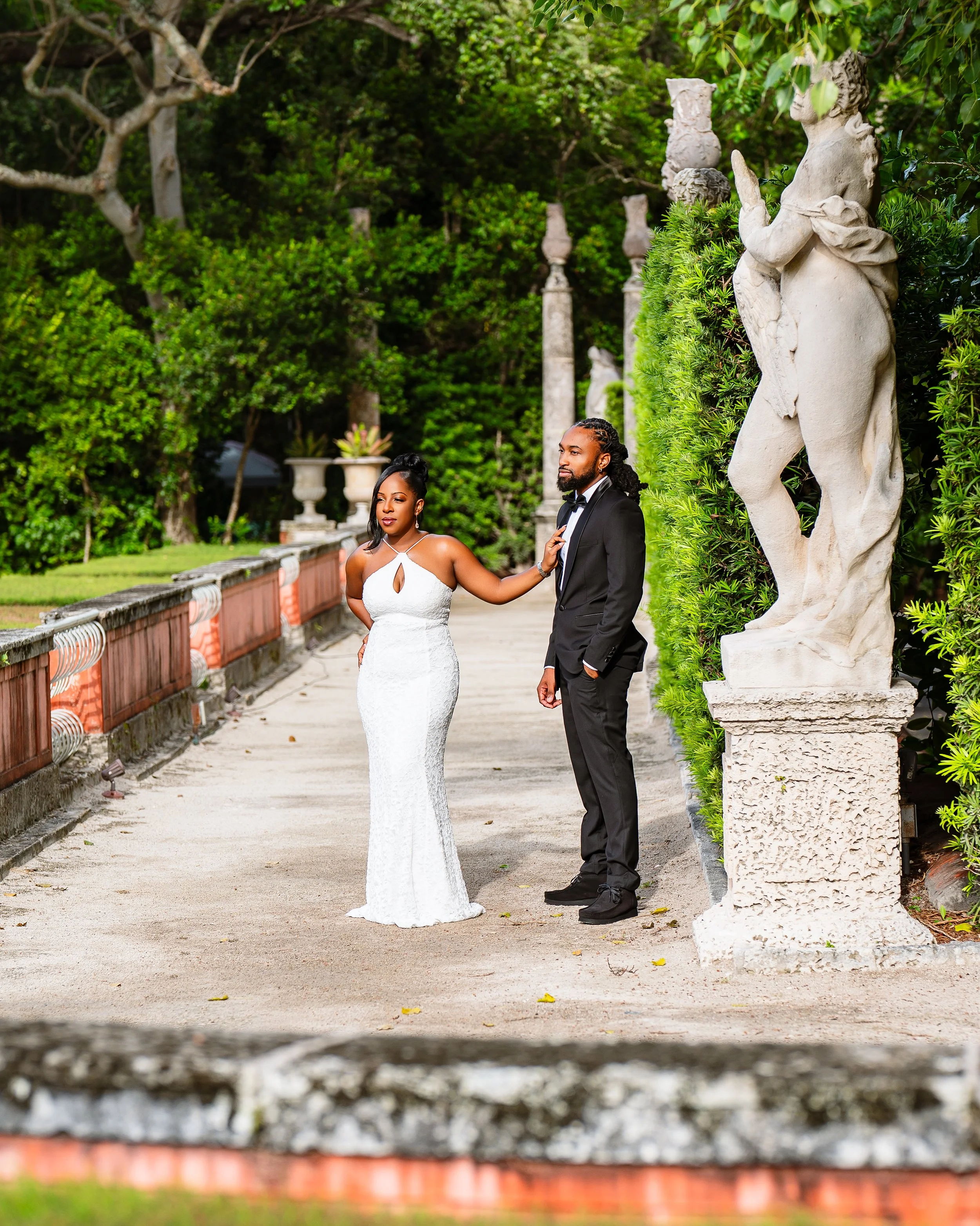 A bride and groom standing outdoors in front of a lush green hedge, with a large stone statue of a woman holding a jug on the right, and a stone-lined road with a red brick wall at the bottom.