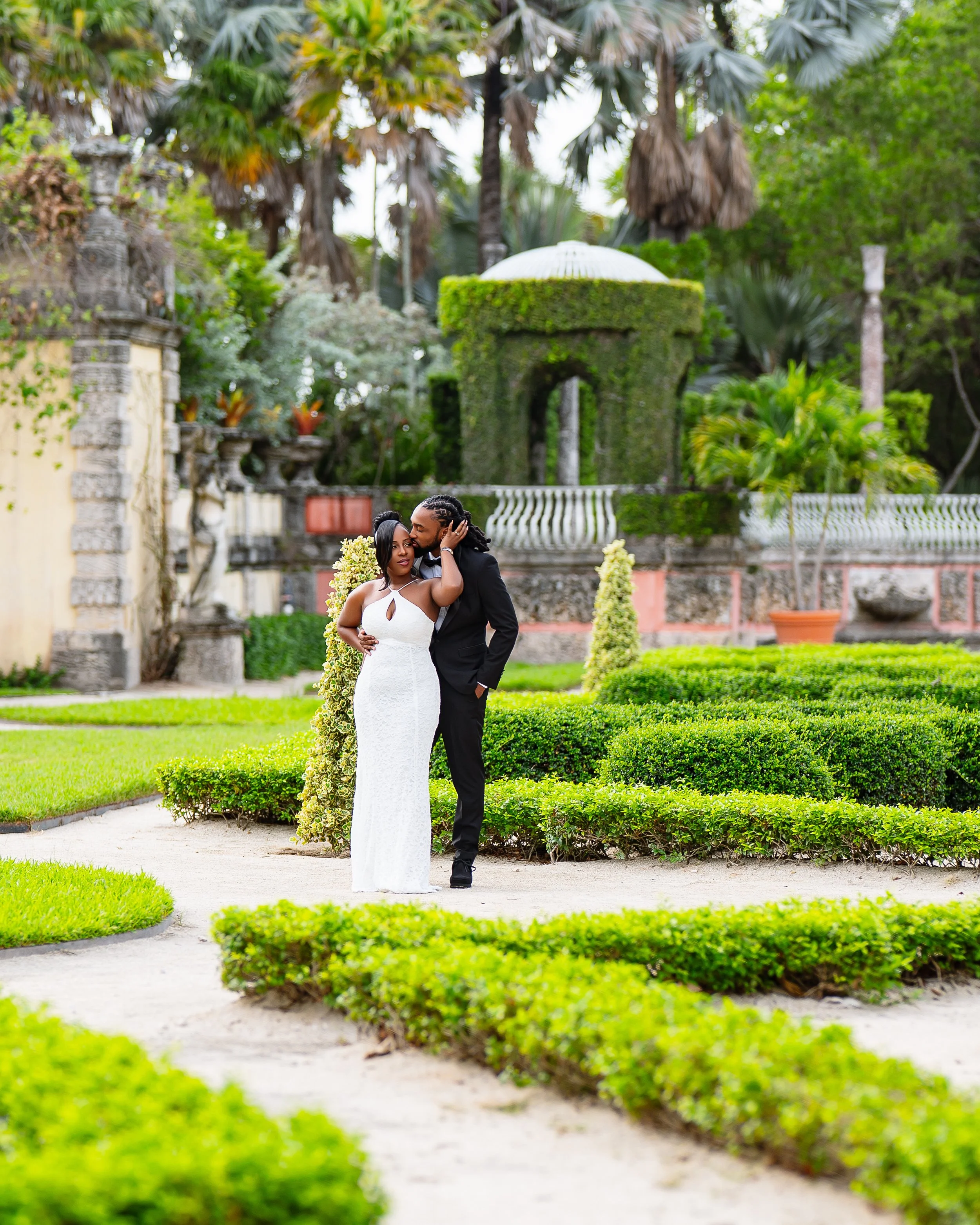 A couple dressed in formal wedding attire hugging in a lush garden with manicured bushes, trees, and a decorative stone gazebo in the background.