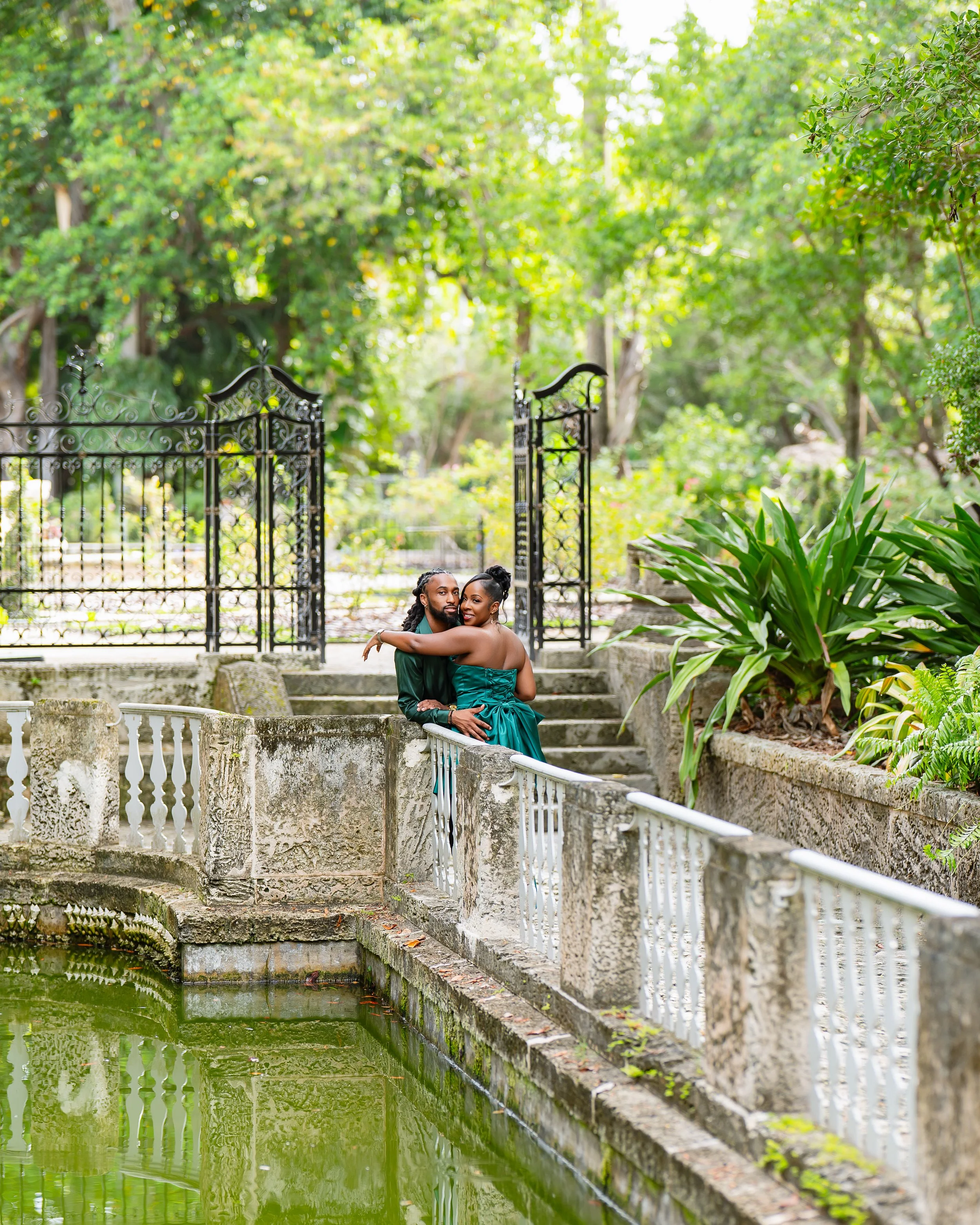 Couple sitting on a stone ledge by a green pond in a lush garden with trees and plants, black wrought-iron gate behind them.