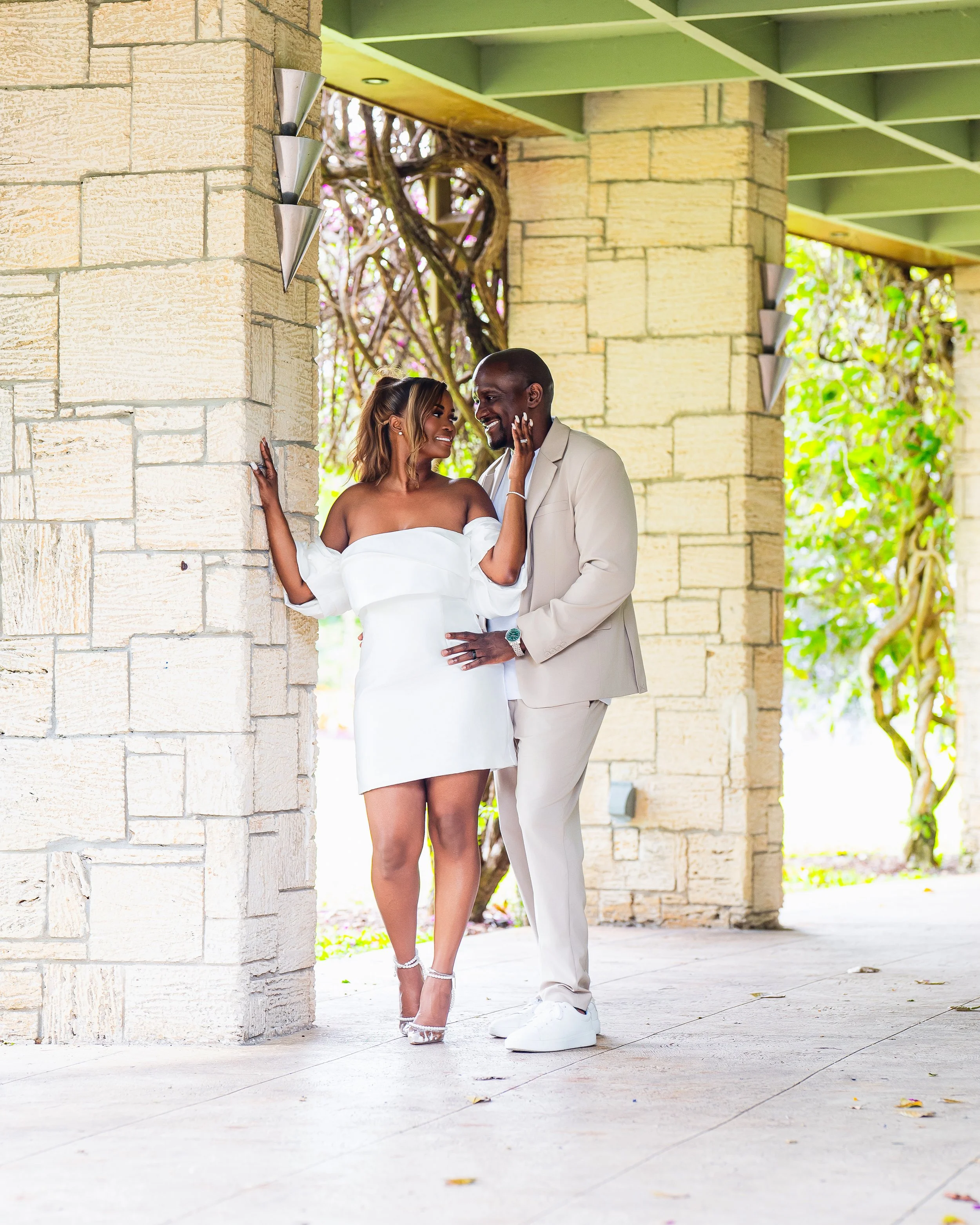 A couple dressed in white poses under a stone archway, smiling and looking at each other. The woman wears an off-the-shoulder dress and high heels, while the man wears a suit and white sneakers. The setting appears to be outdoors with greenery in the