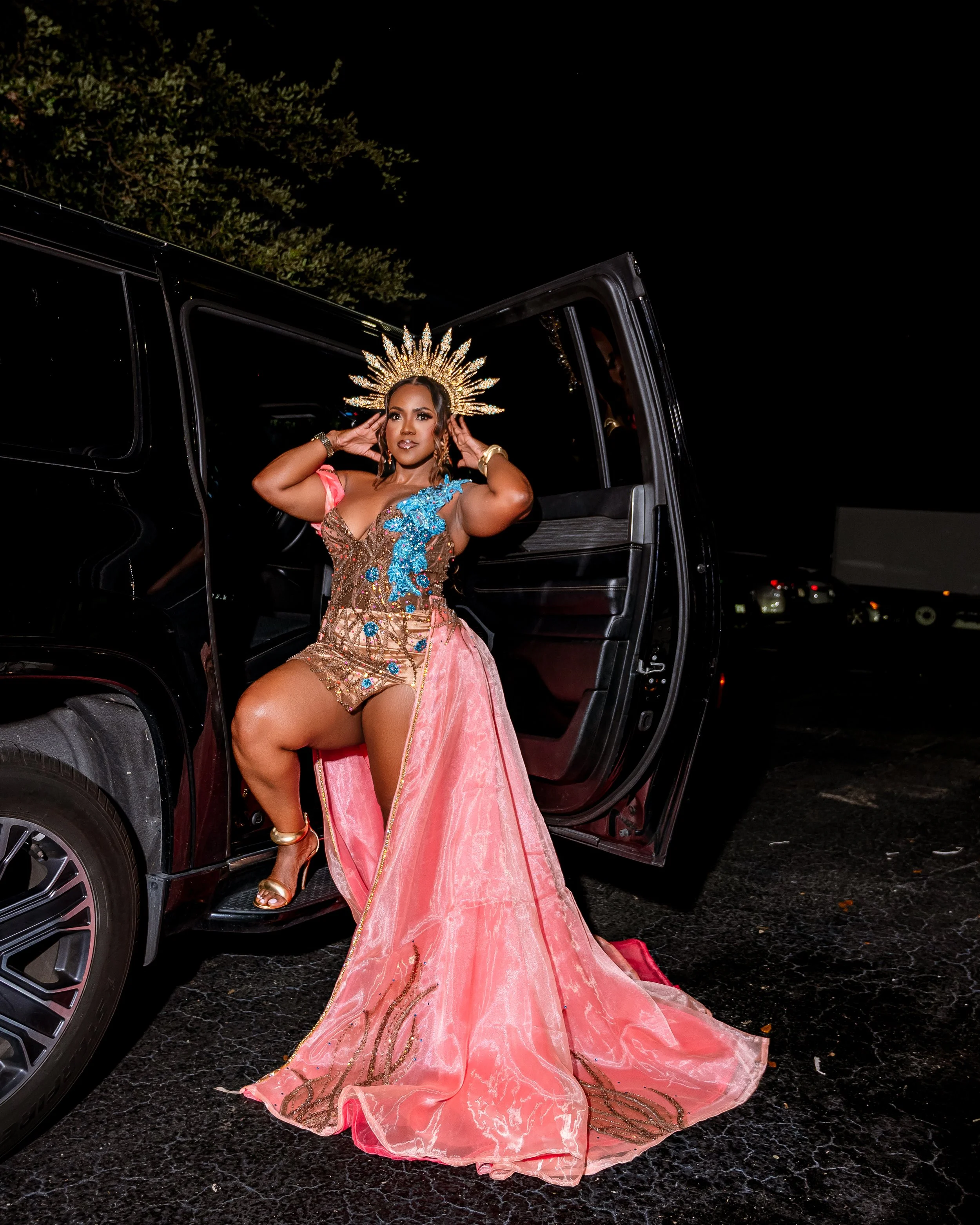 A woman dressed in an elaborate, colorful gown with intricate blue and gold embellishments, stepping out of a black vehicle at night. She is wearing a large, ornate halo-like headpiece, gold jewelry, and high heels, and is holding her head with both 