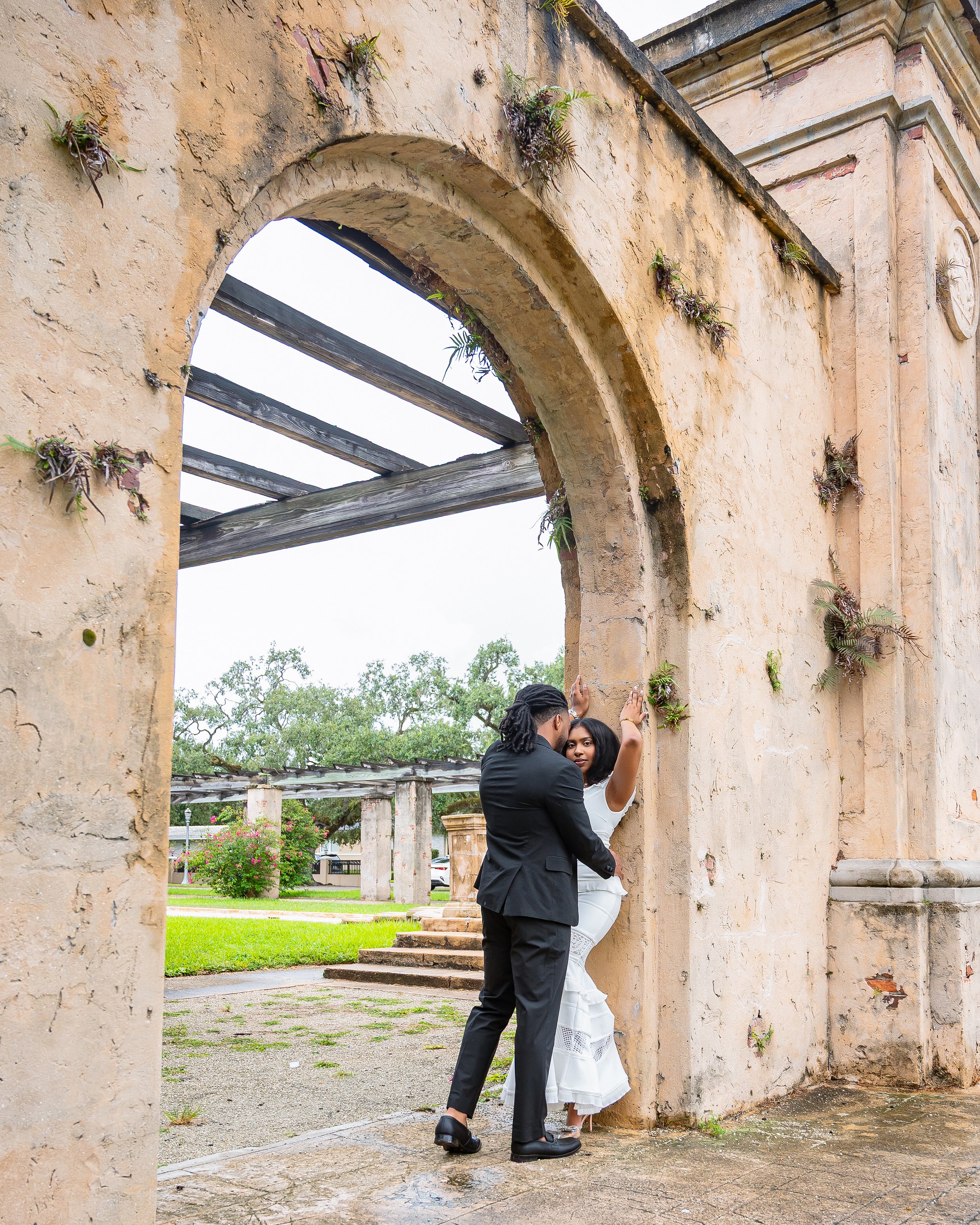A couple dressed in formal attire shares a kiss underneath a stone archway. The man is wearing a black suit, and the woman is dressed in a white dress. The scene takes place outdoors, with greenery and flowers visible in the background.