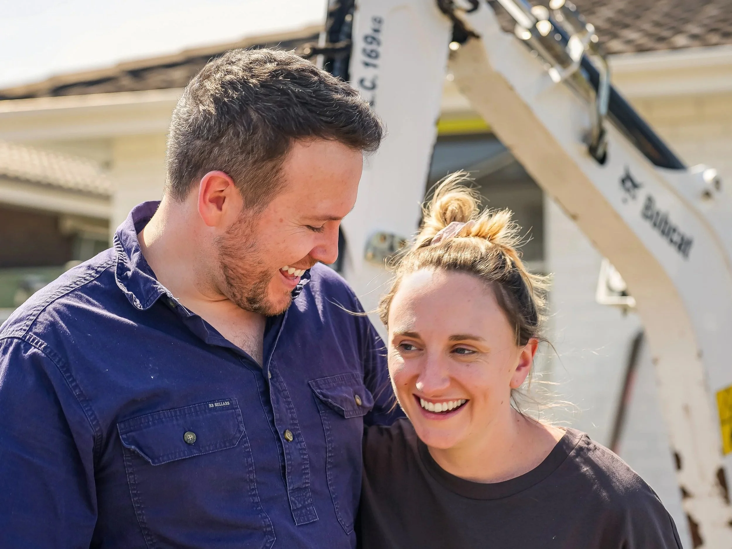 A smiling couple, Amelia and James, on the site of a renovation.