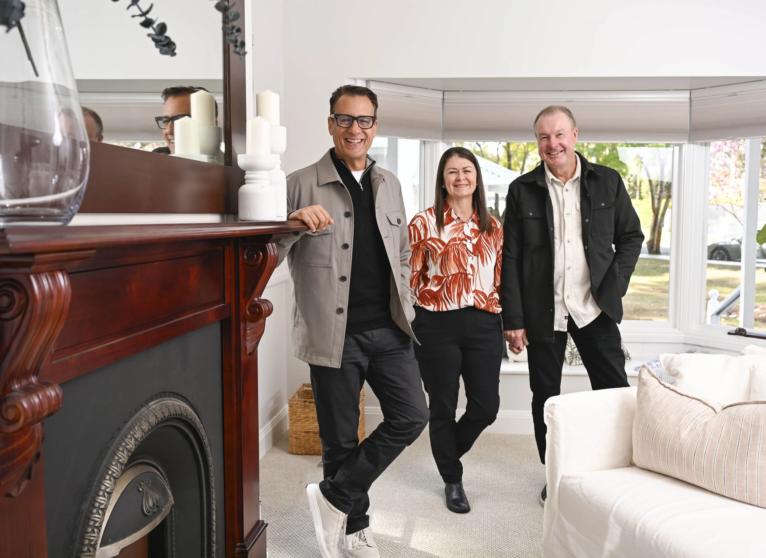 Selling Houses Australia host Andrew Winter with homeowners Jenny and Steve, standing smiling by the living room fireplace, post renovation.