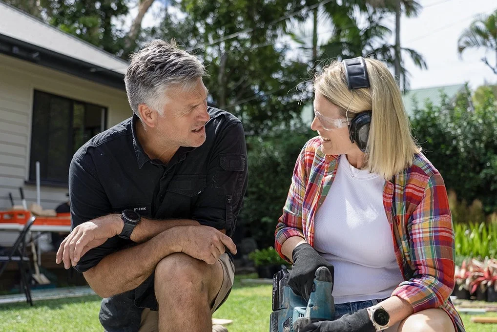 Landscaper Dennis Scott and interior designer Wendy Moore looking at one another in a garden. Wendy is wearing safety gear and holding a drill.