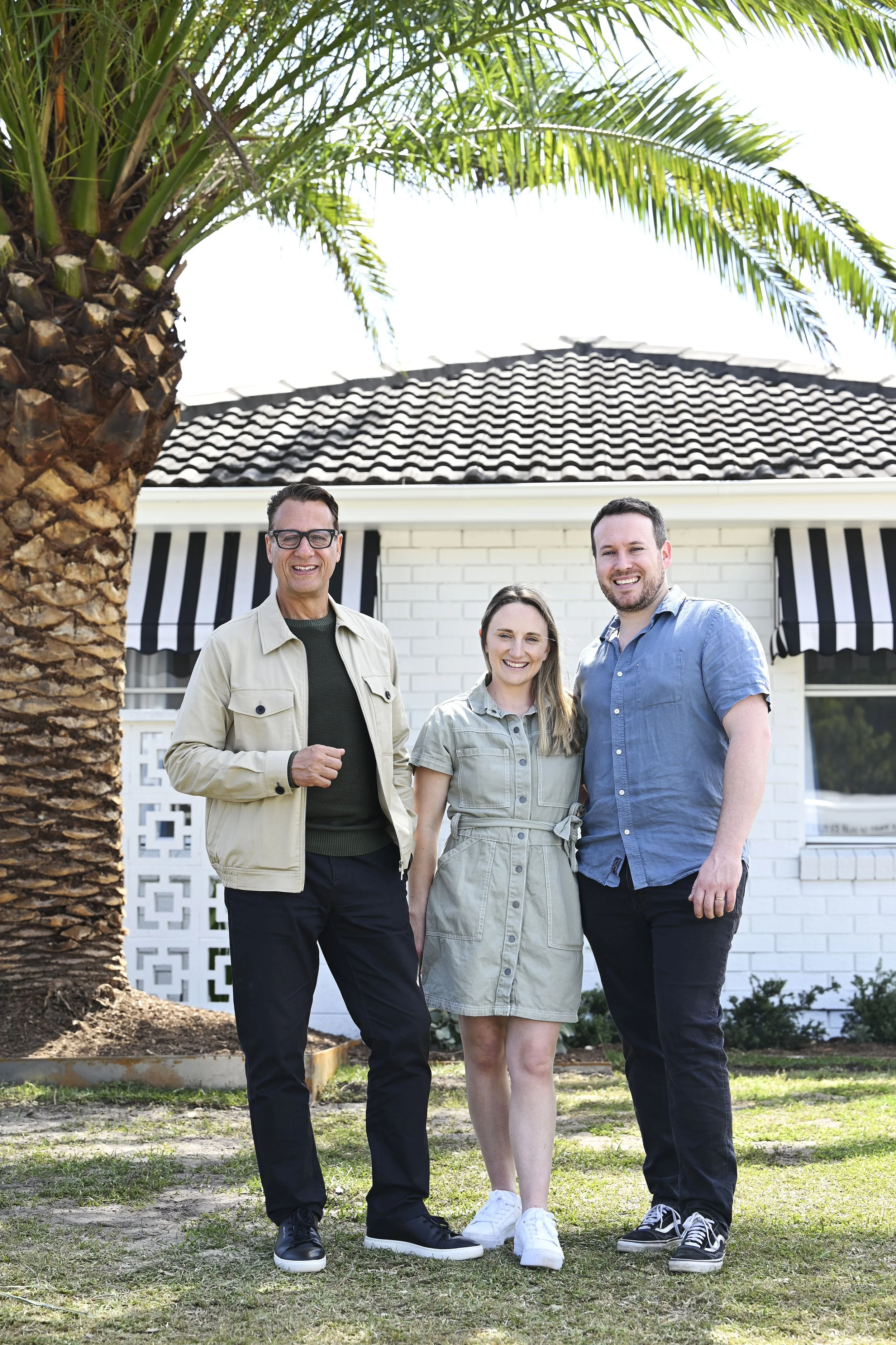 Selling Houses Australia host Andrew Winter and homeowners Amelia and James smile, standing in front of a newly painted white brick house and large palm tree.