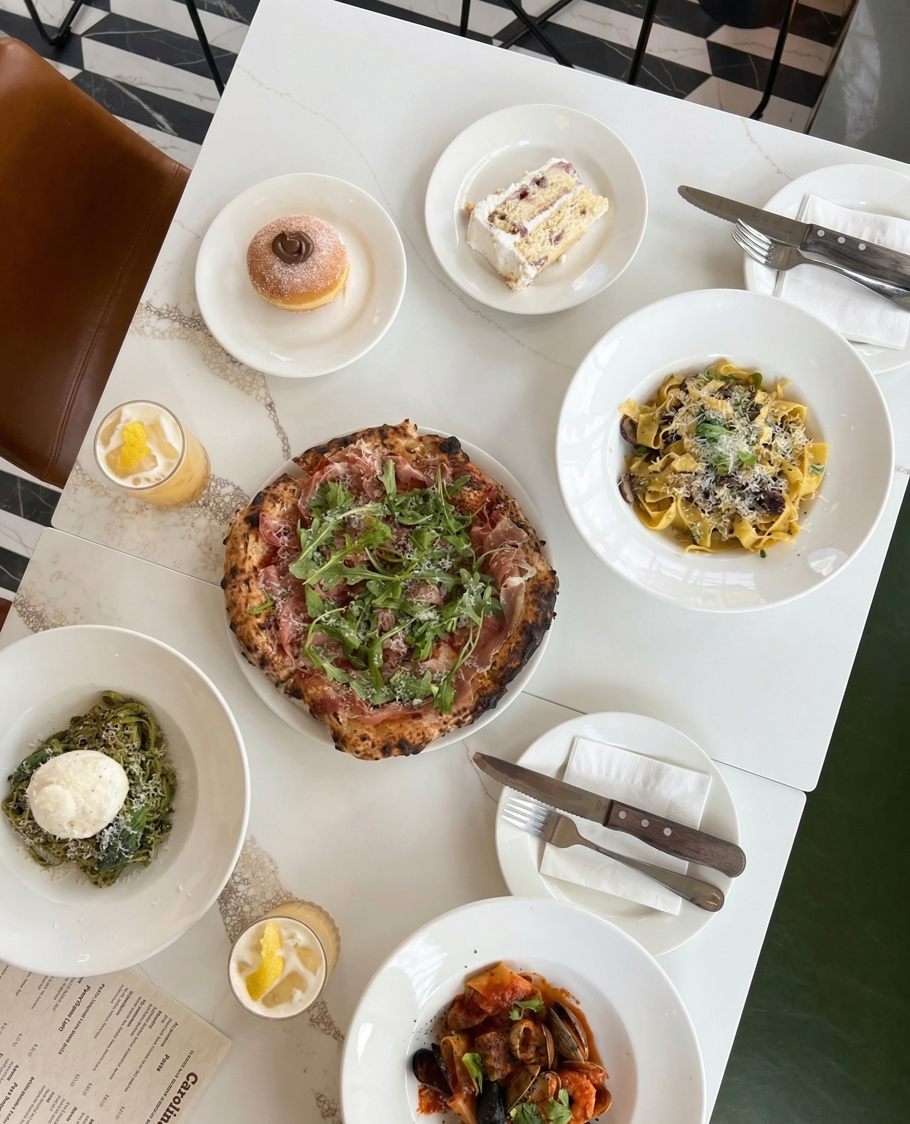 Table set with various dishes including pizza, pasta, and desserts, along with drinks and cutlery.
