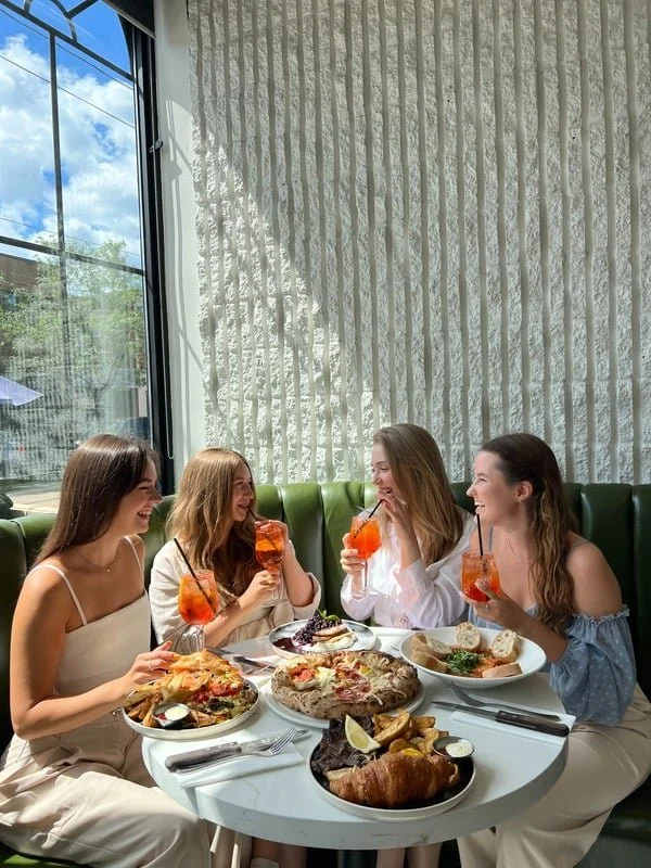 Four women sitting at a round table in a restaurant, enjoying pizza and drinks, with a window showing a clear sky outside.