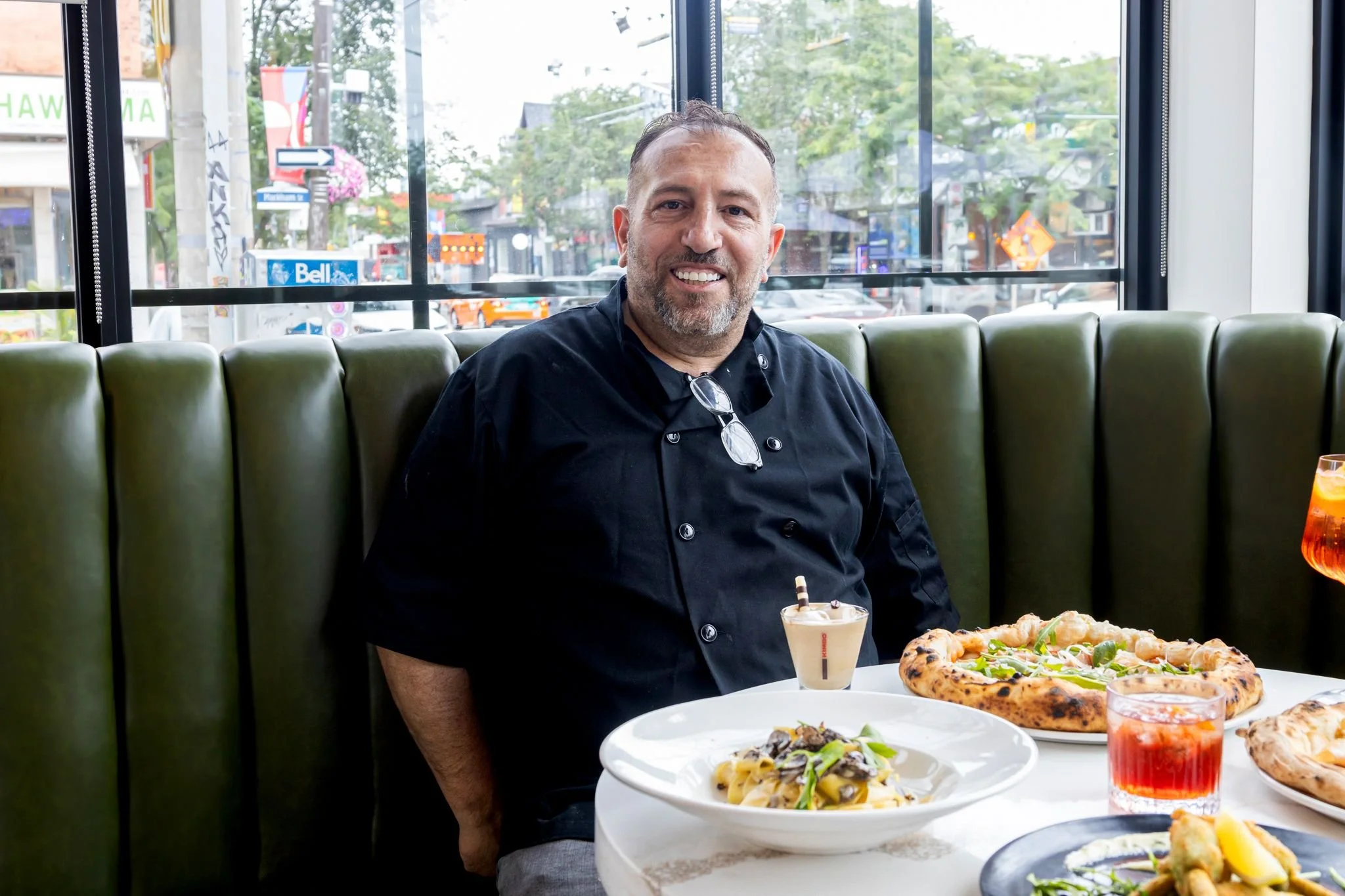 A man with short hair and a beard smiling while sitting at a restaurant table with pizza and pasta.