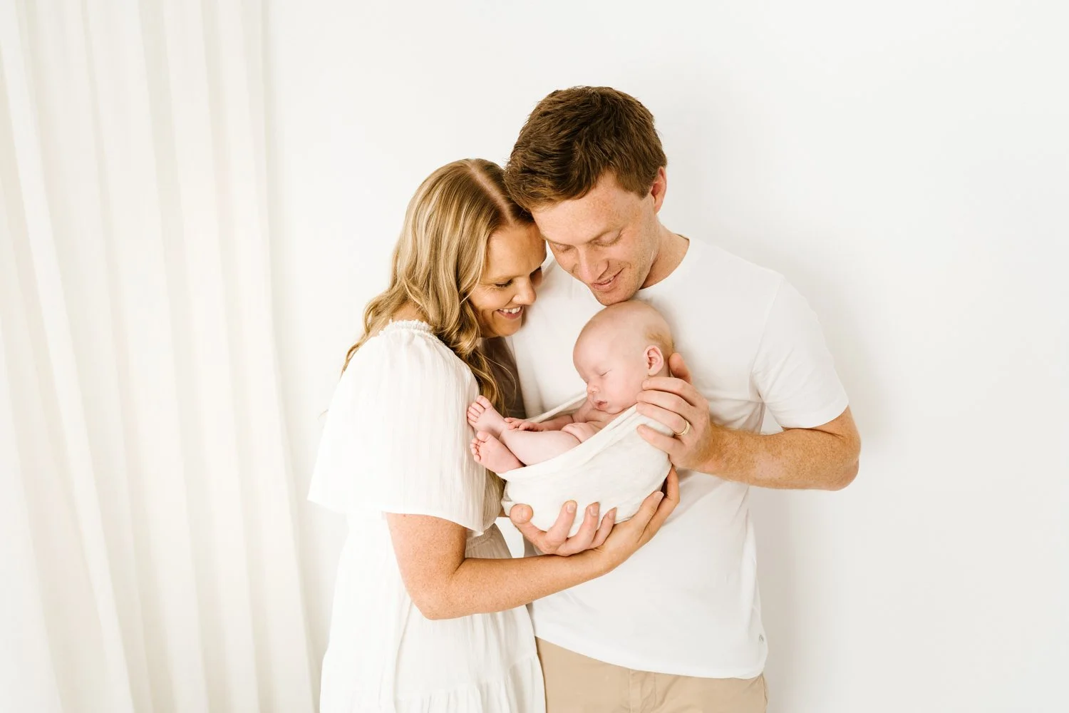A happy couple holding their newborn baby together in a bright, white room.