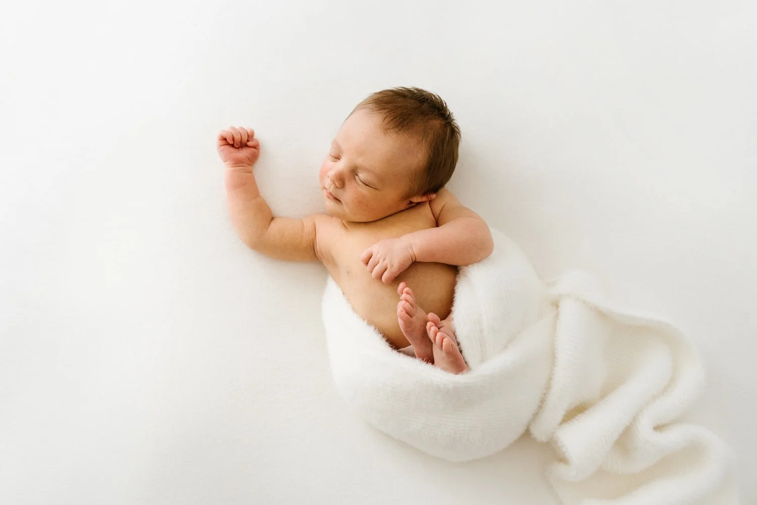 Newborn baby sleeping on a soft white surface wrapped partially in a white blanket, with a peaceful expression and tiny clenched fists.