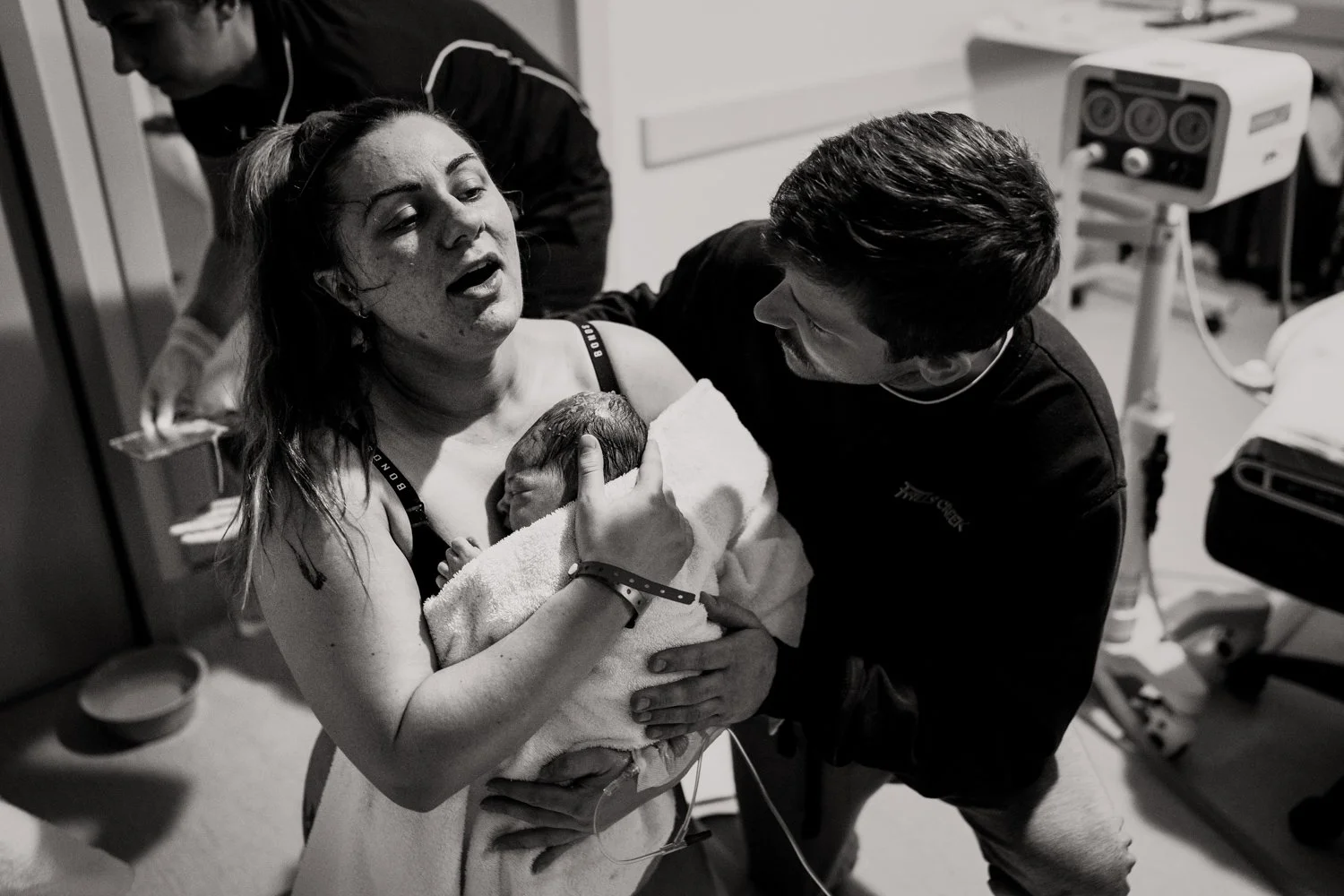 A woman in a hospital bed holding a newborn baby, smiling at the baby, with a man sitting nearby, smiling back in a hospital room with medical equipment.