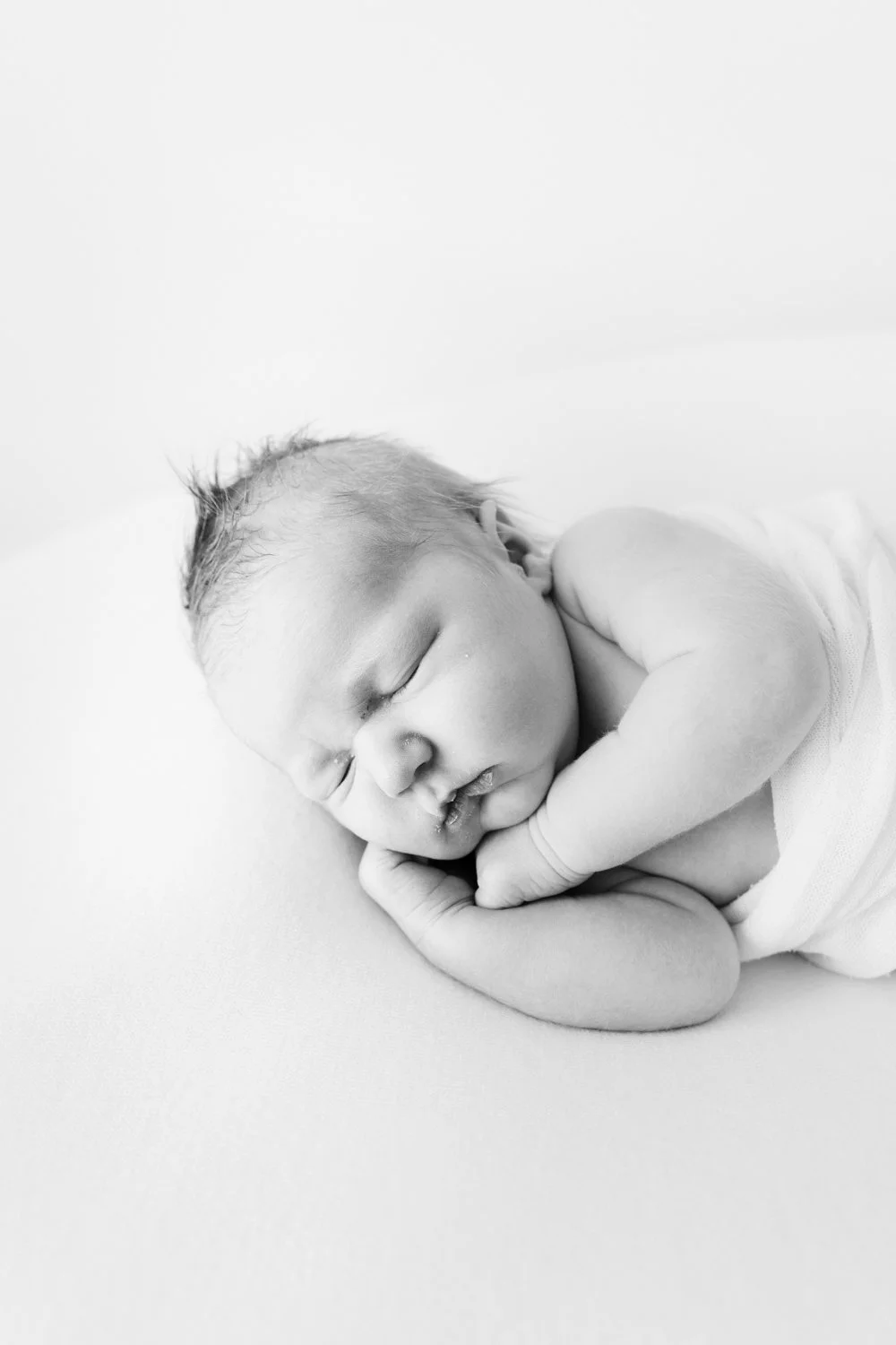 Black and white photo of a peacefully sleeping newborn baby, curled up with hands near face, on a soft surface.