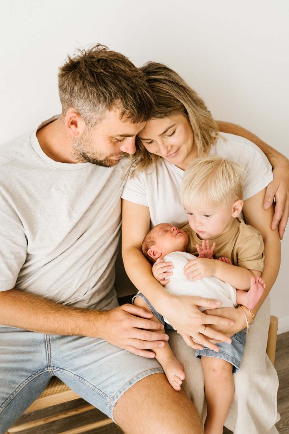Family of four, including a man, woman, and two young children, sitting together and holding a newborn baby. They are smiling and gazing lovingly at the baby.