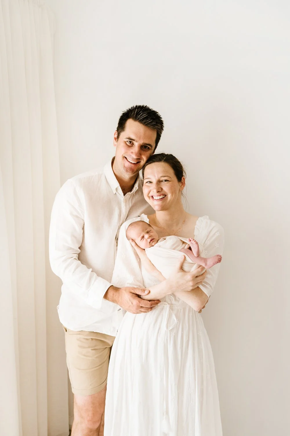 A smiling family of three with a newborn baby, all dressed in white, standing against a plain white background.