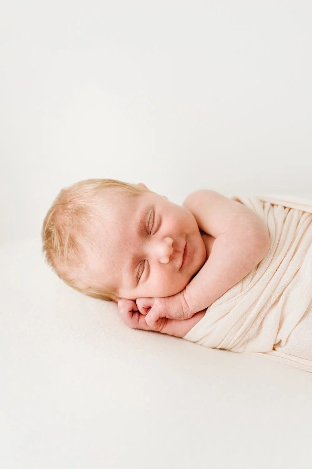 A sleeping baby with blonde hair, resting on a white surface, wrapped in a light-colored blanket, with a peaceful expression.