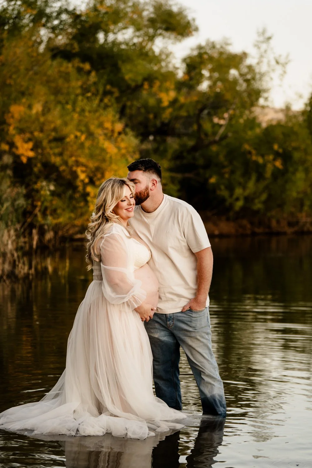 A man and a pregnant woman standing in a body of water during sunset, with trees and autumn leaves in the background. The woman is wearing a flowing white dress, and the man is dressed in a light-colored shirt and jeans. They are close together, smil