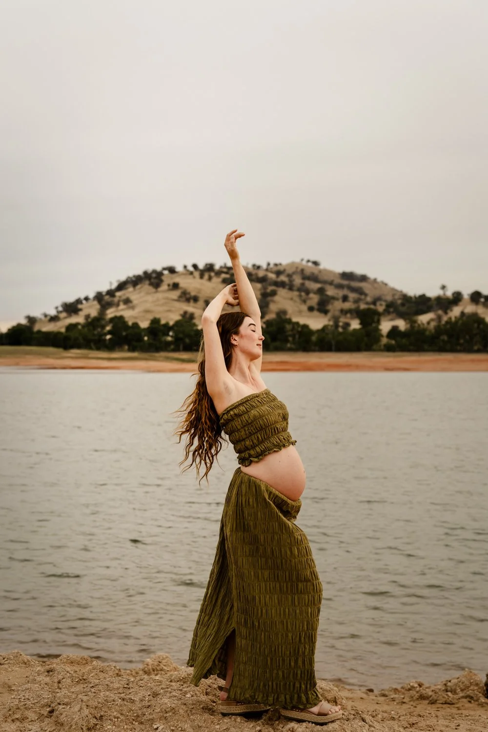 A pregnant woman with long curly hair stands on a rocky shore by a lake, wearing a matching green strapless top and long skirt, with her arms raised above her head, eyes closed, and a mountain with sparse trees is visible in the background.