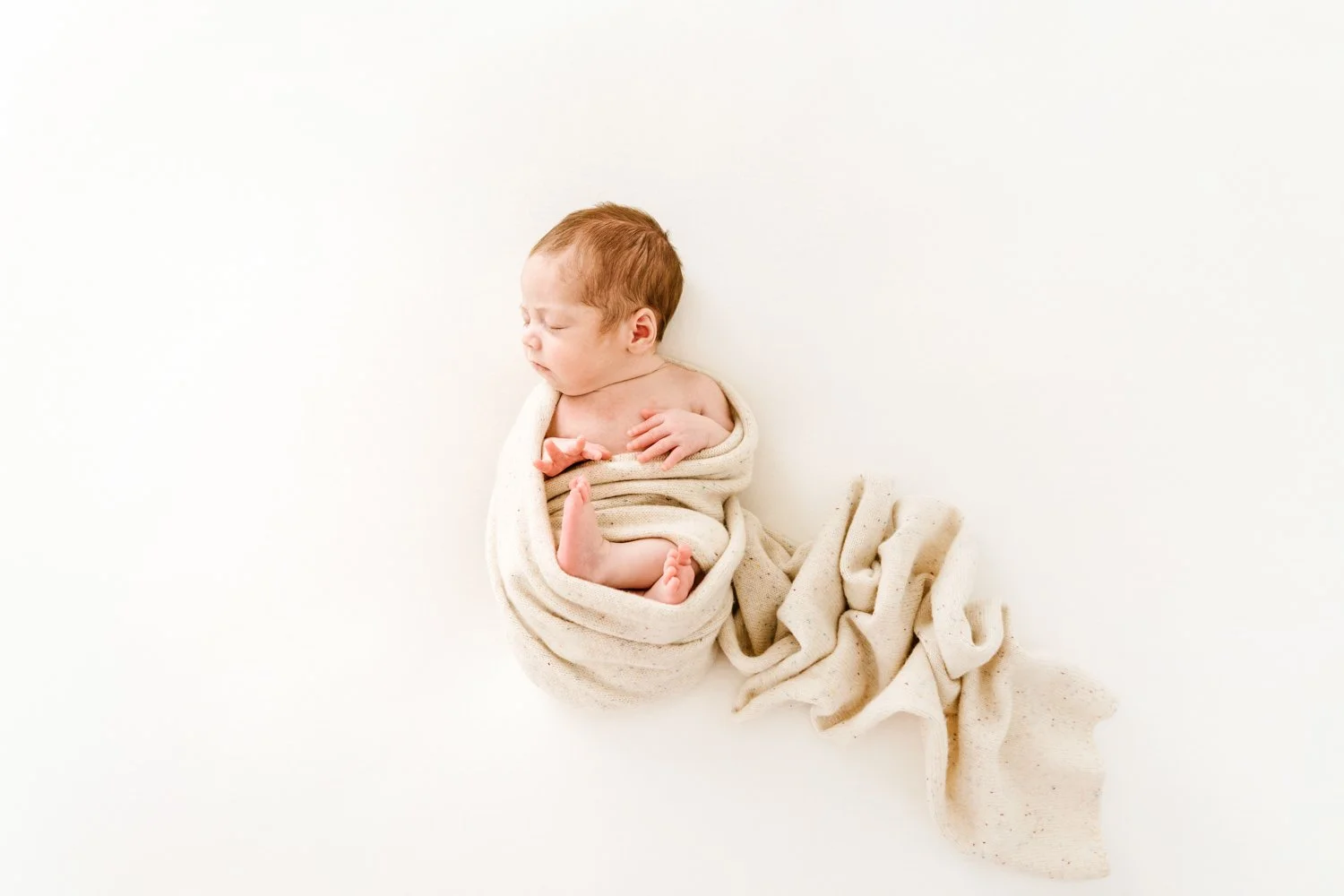 A sleeping newborn baby with reddish hair, wrapped in a cream-colored blanket, lying on a white background.