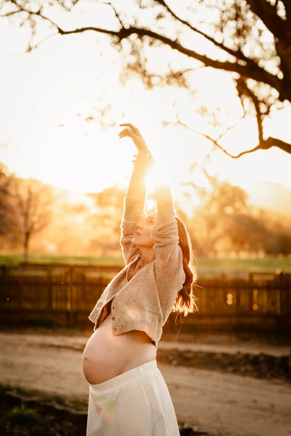 Pregnant woman outdoors at sunset, stretching her arms upward, with trees and a fence in the background.
