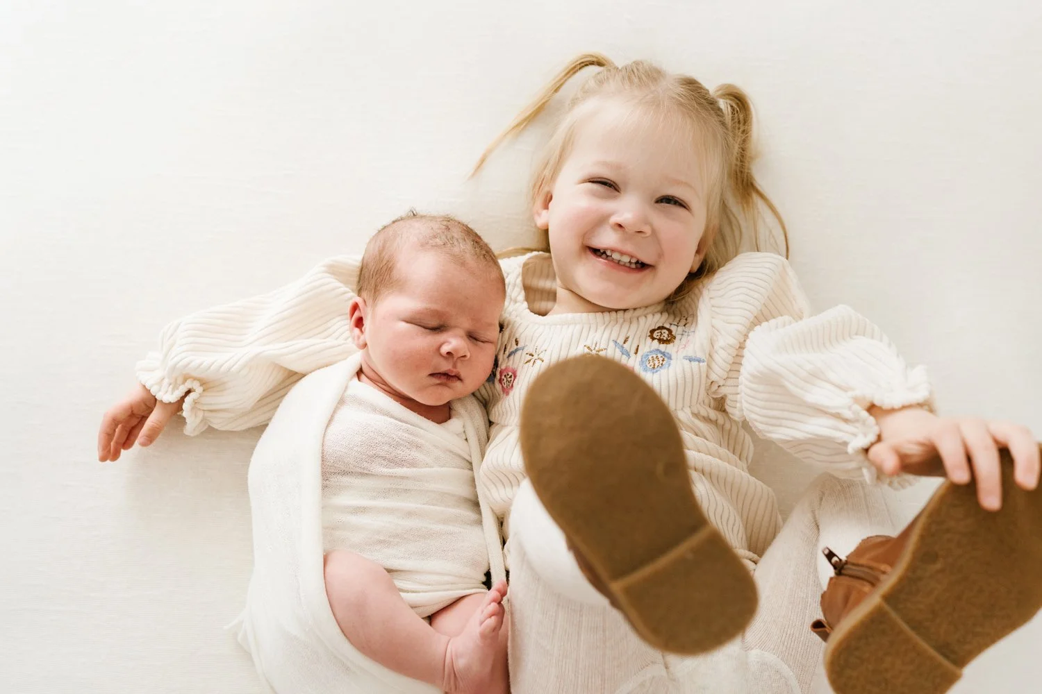 A young girl with blonde hair and a big smile lying on her back, holding a sleeping baby wrapped in a light blanket. The girl is wearing a cream-colored sweater with embroidered flowers and has her legs raised, showing brown shoes.