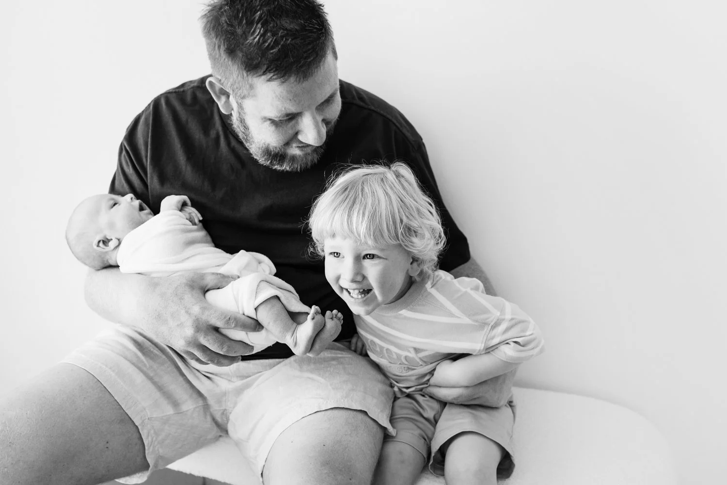 A man holding a newborn baby while a young boy sitting next to them smiles and leans toward the baby.