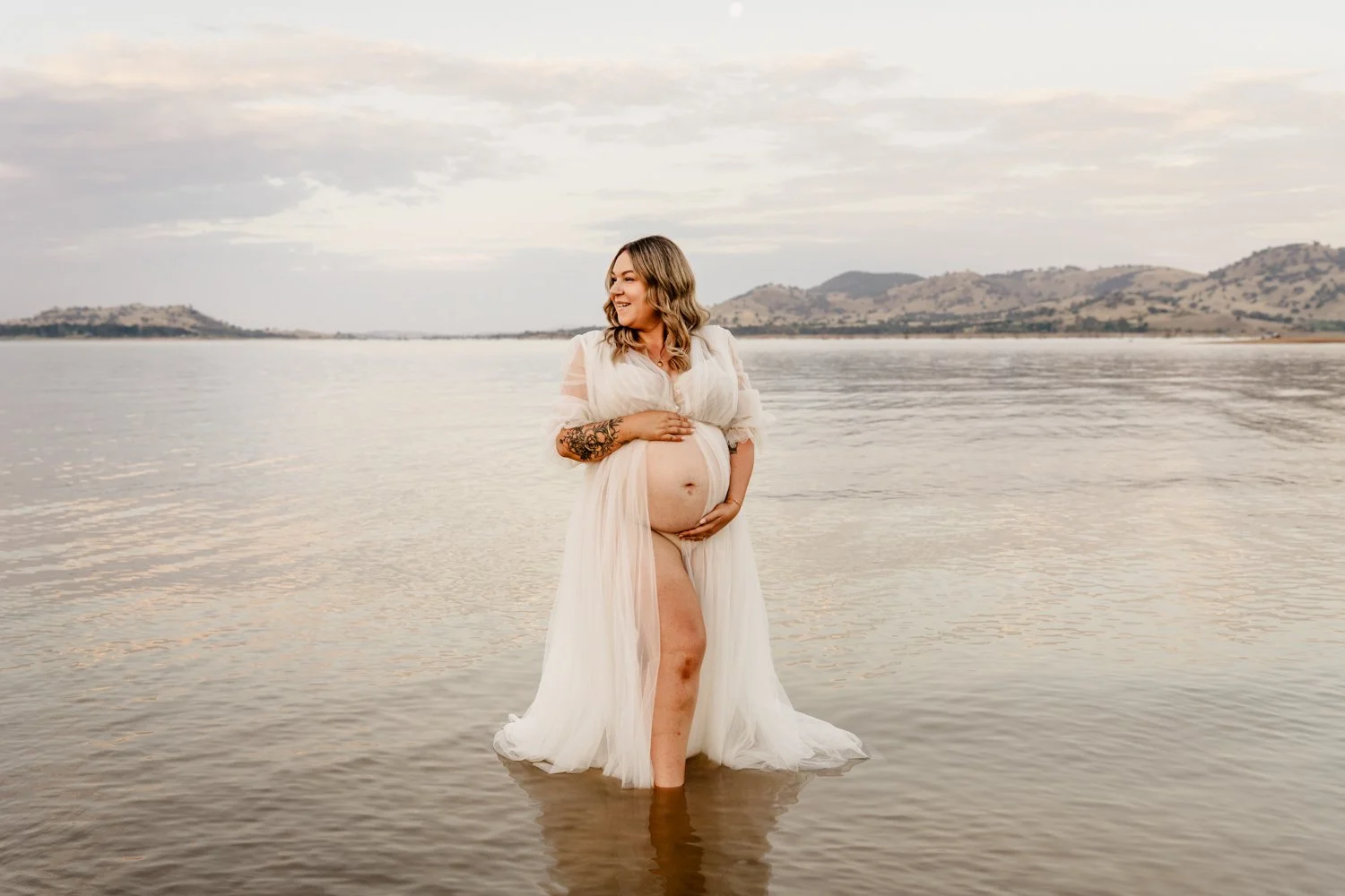 A pregnant woman standing in shallow water at the beach, wearing a flowing light-colored dress, with a scenic backdrop of distant mountains and a cloudy sky.