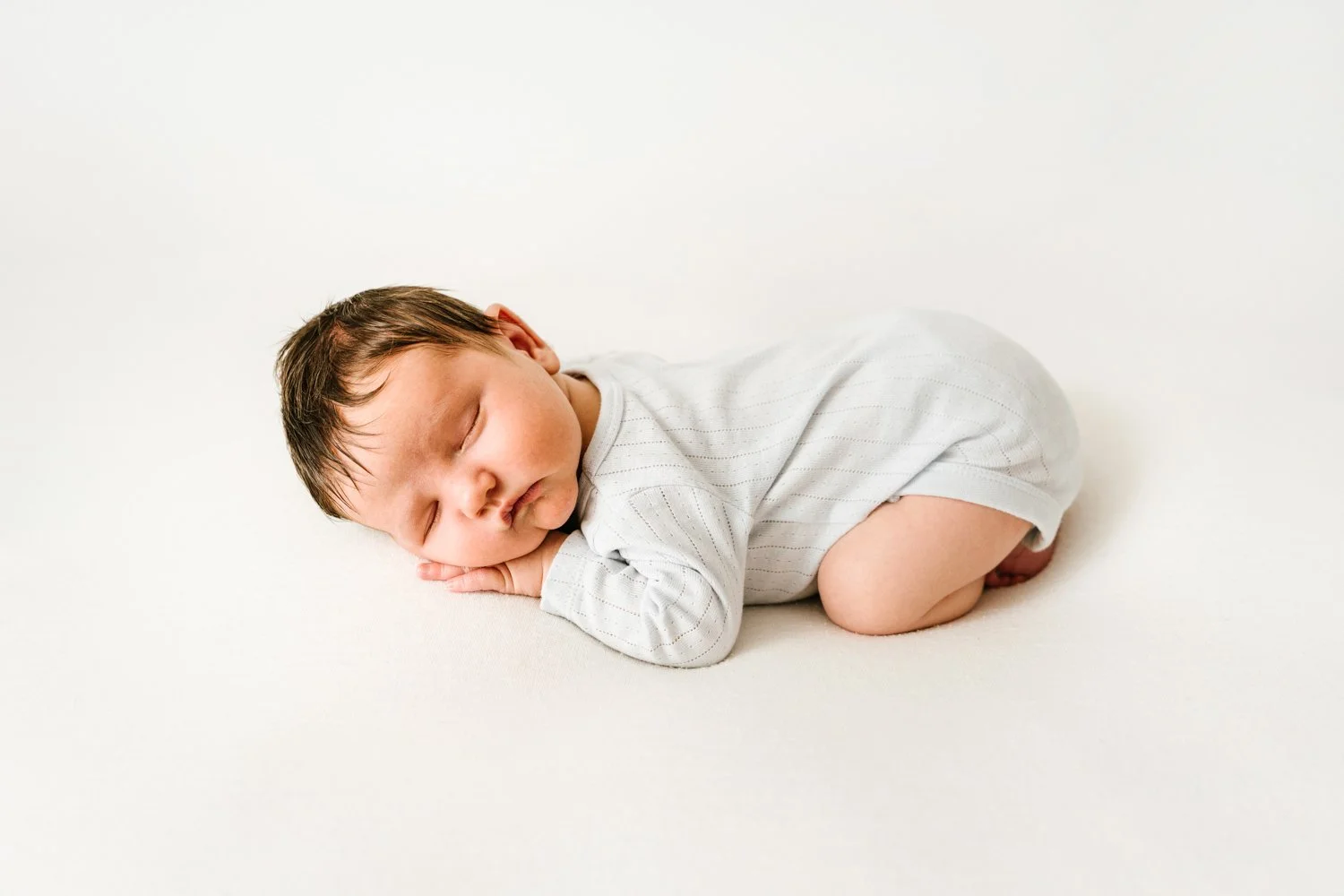 A sleeping baby lying on its side with hands under face, on a white surface, wearing a white long-sleeve shirt.