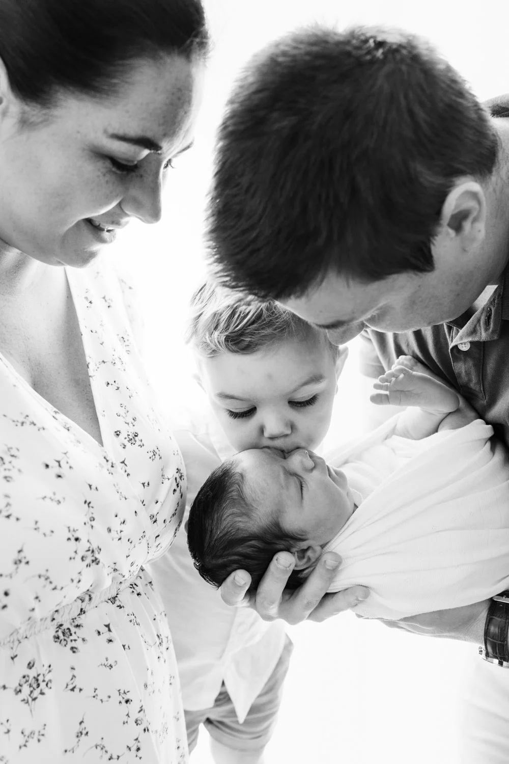 Family gathered around newborn baby, kissing newborn in a black and white photo.