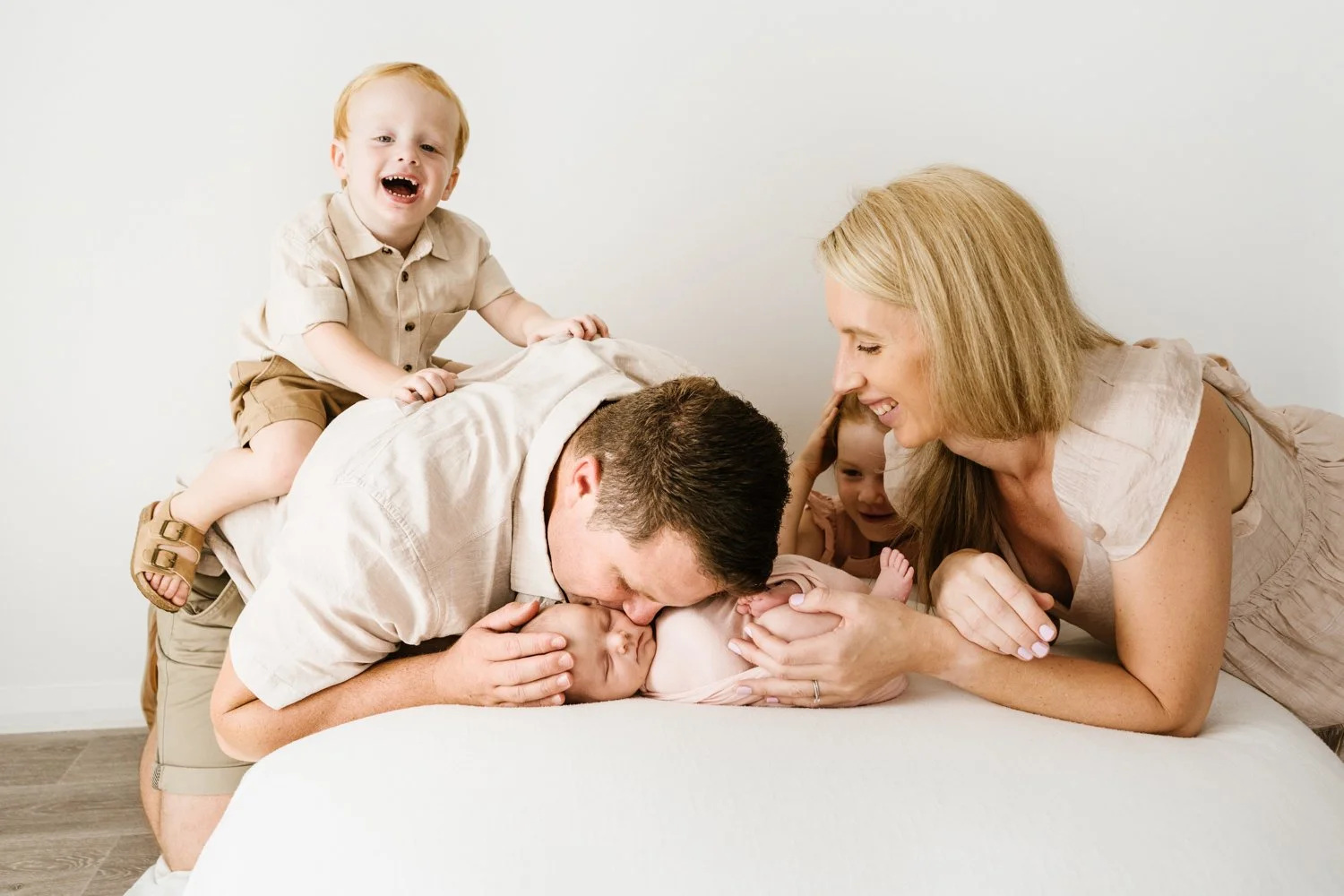 Family of four celebrating a newborn, with a father kissing the baby on the forehead, mother smiling at the baby, and two young children on the bed.