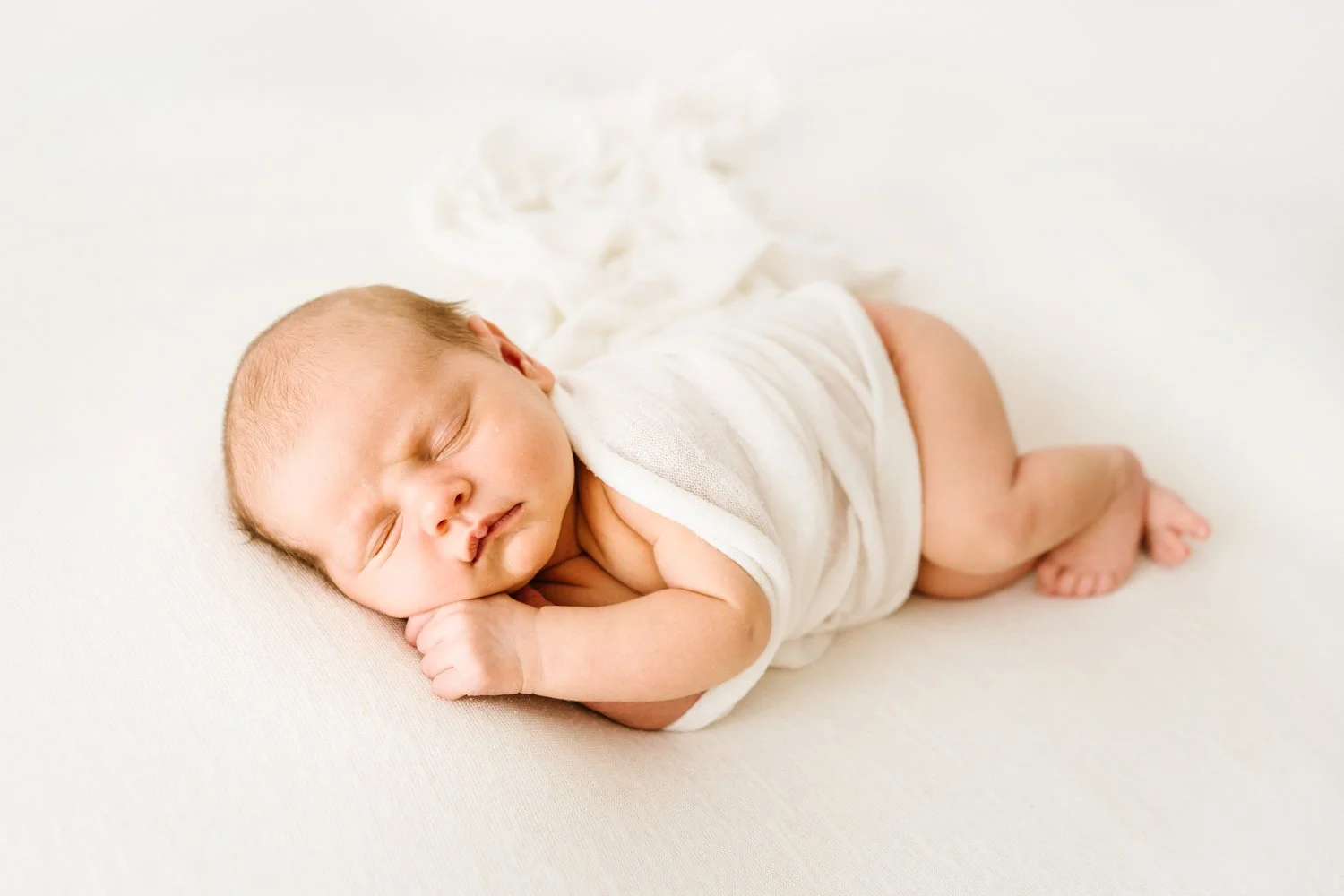 A sleeping newborn baby lying on a soft surface, wrapped in a white cloth, with arms curled up near the face and eyes closed.