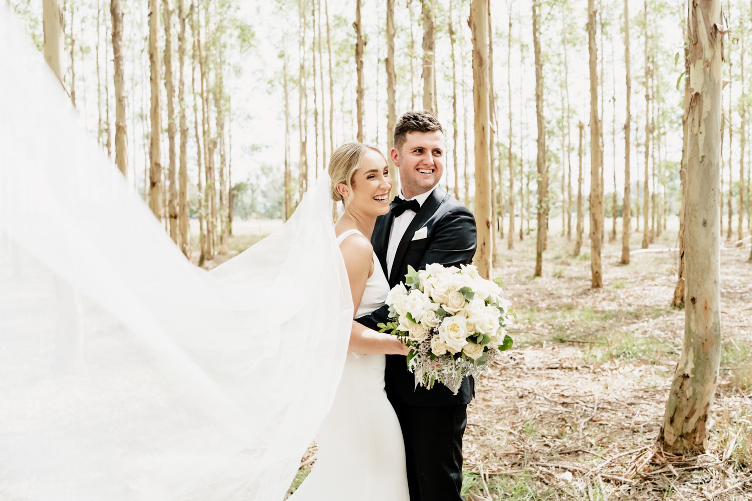 portrait of bride and groom in wedding attire. Bride holding bouquet amongst the trees