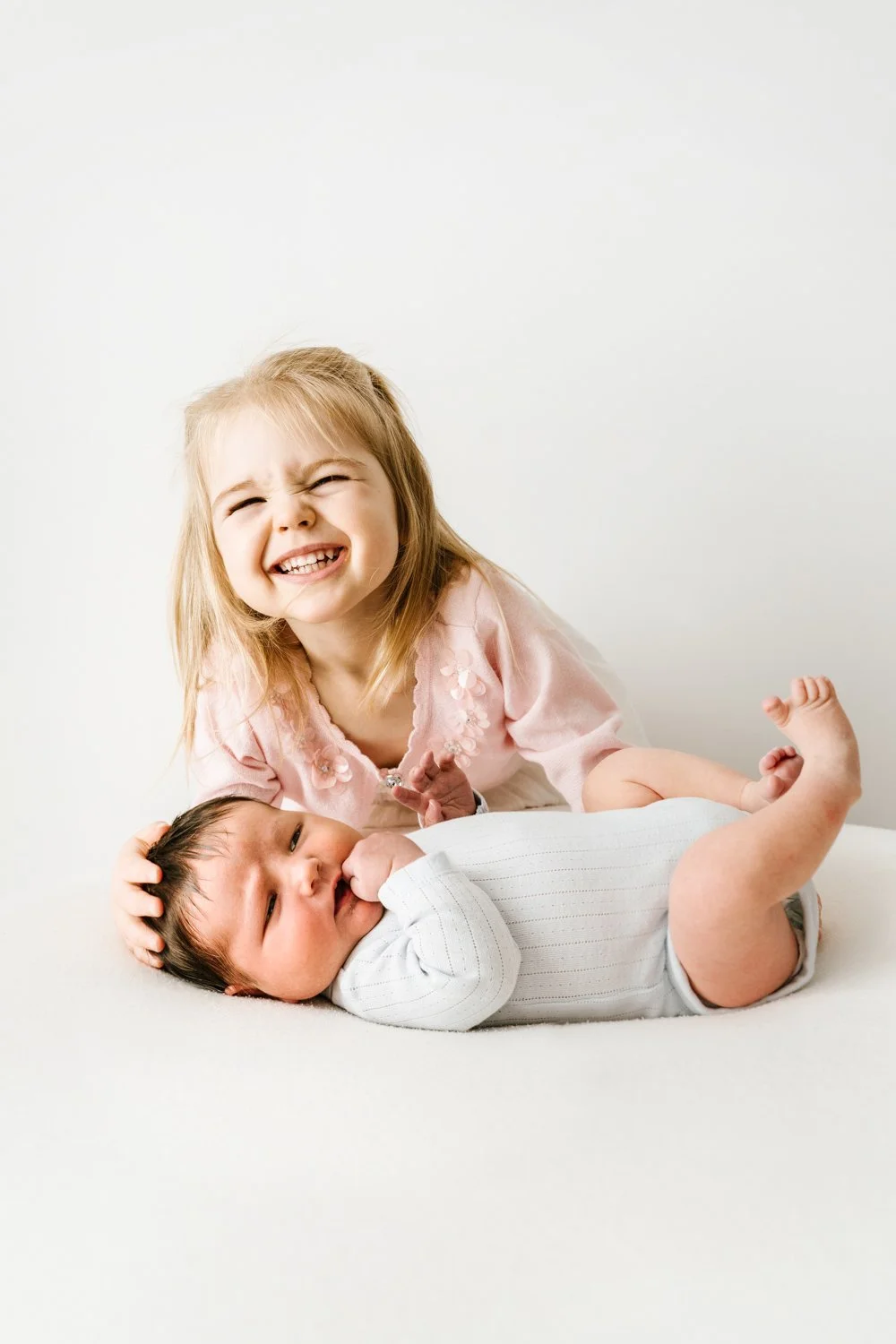 A young girl with blonde hair smiling and playing with a baby lying on a white surface.