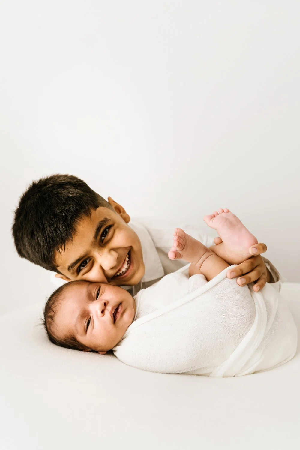 A young boy with short dark hair smiling and hugging a newborn baby wrapped in a white blanket.
