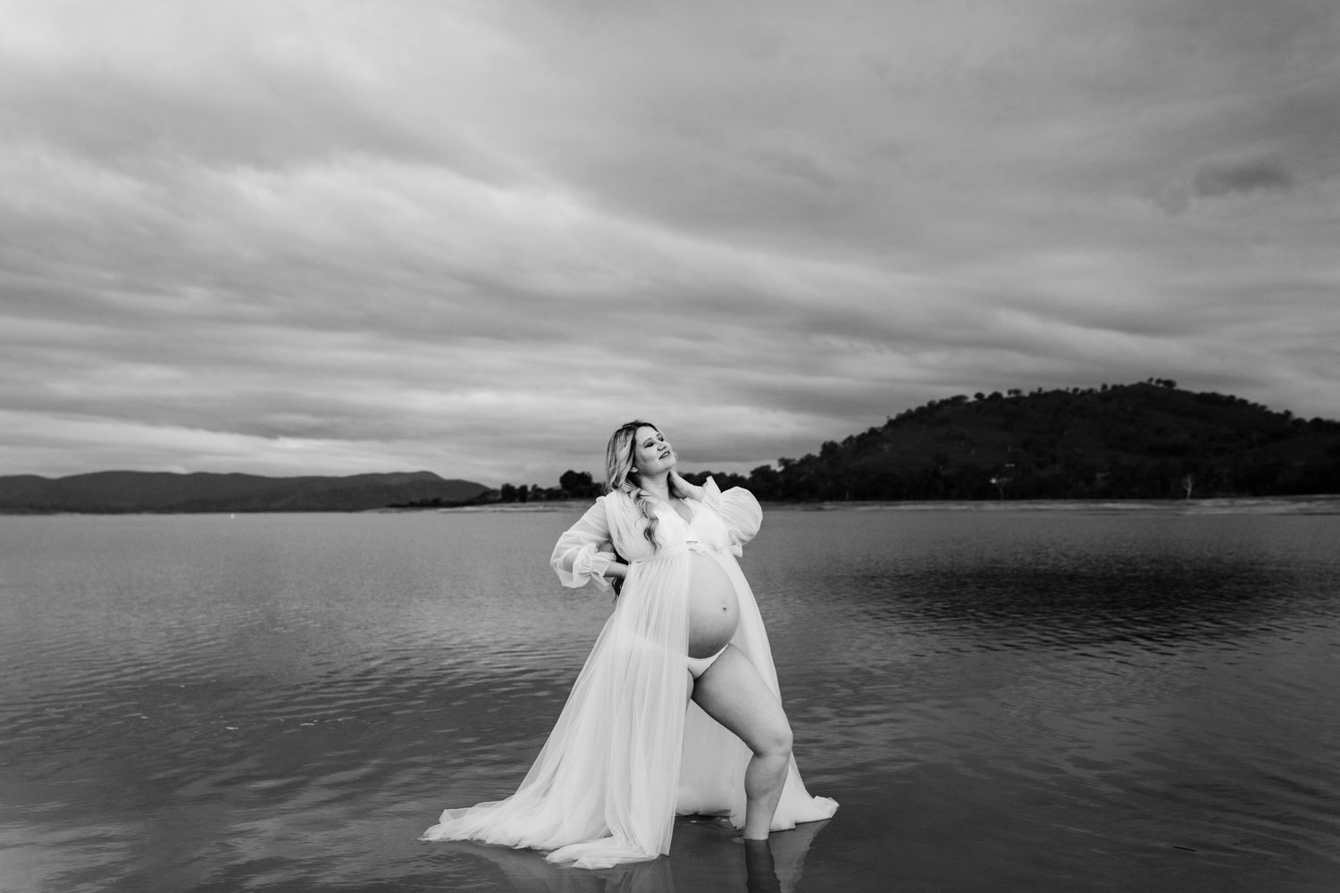 A pregnant woman in a flowing dress stands in shallow water by a lake, with mountains and cloudy sky in the background.