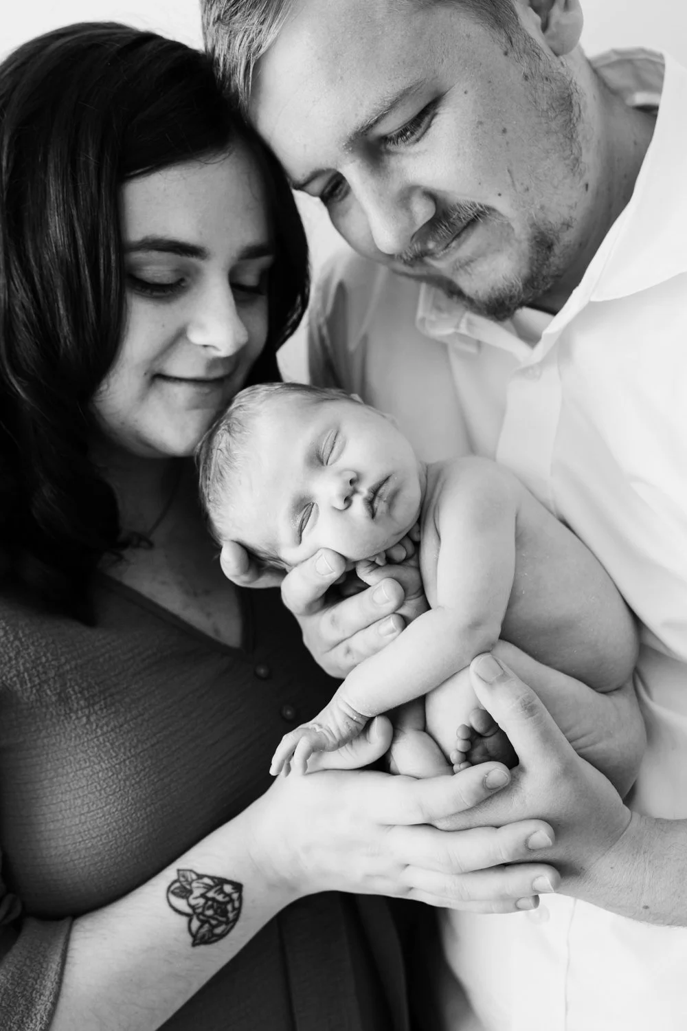 A family of three, with a mother, father, and their newborn baby, close together holding and cuddling the sleeping infant.