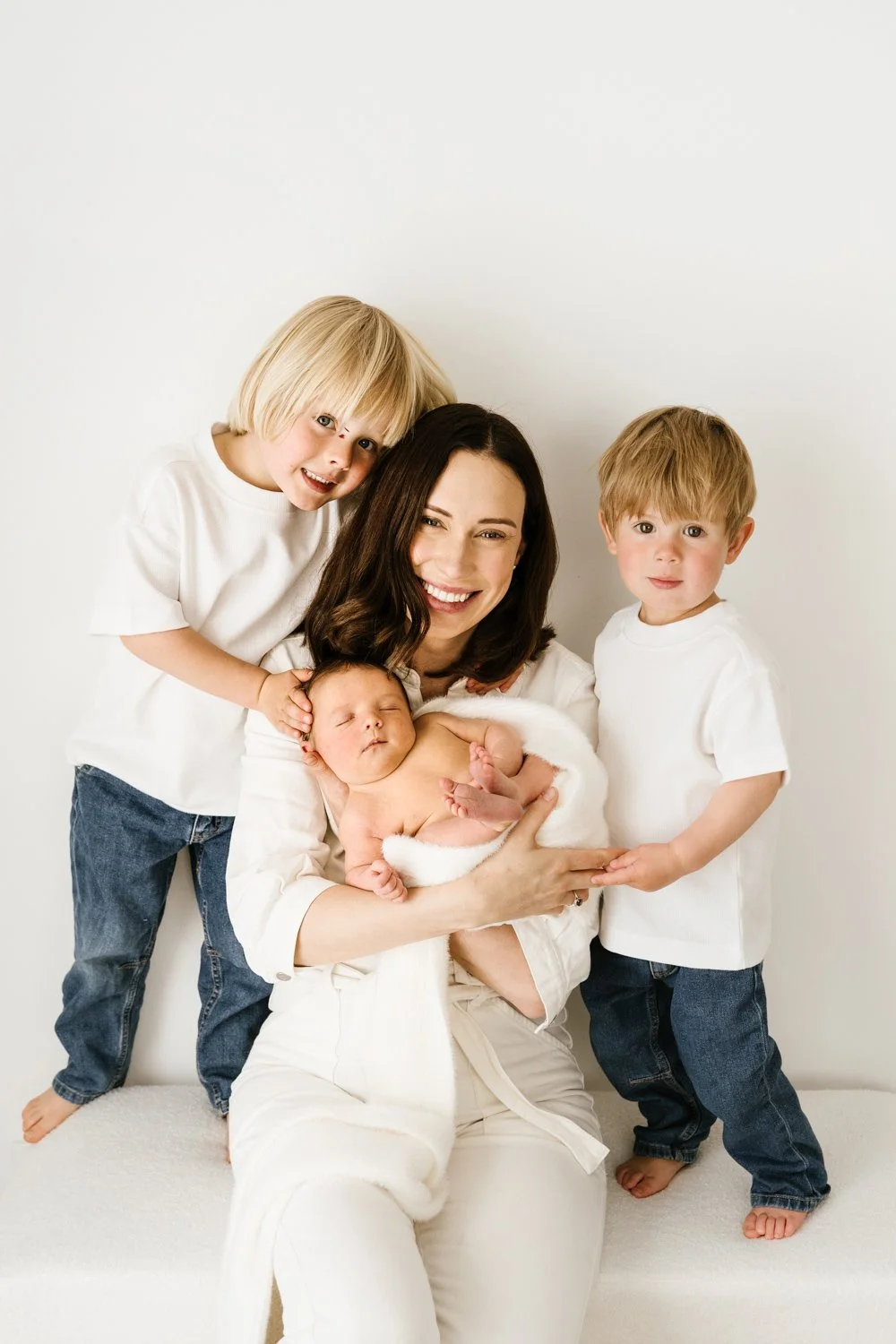 A smiling woman holding a newborn baby, with two young children standing besideher, all dressed in white, against a plain white background.
