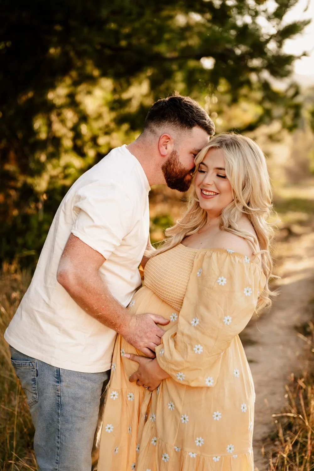 A couple, a man and a pregnant woman, sharing an affectionate moment outdoors at sunset with trees in the background. The man is kissing the woman's forehead while she smiles, holding her belly.