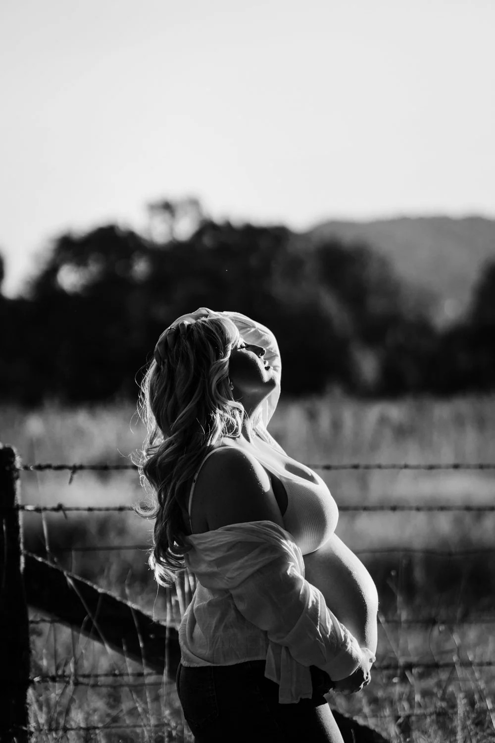 A pregnant woman standing outdoors near a barbed wire fence, with wind blowing her blonde hair, in a black and white photo taken during sunset or sunrise.