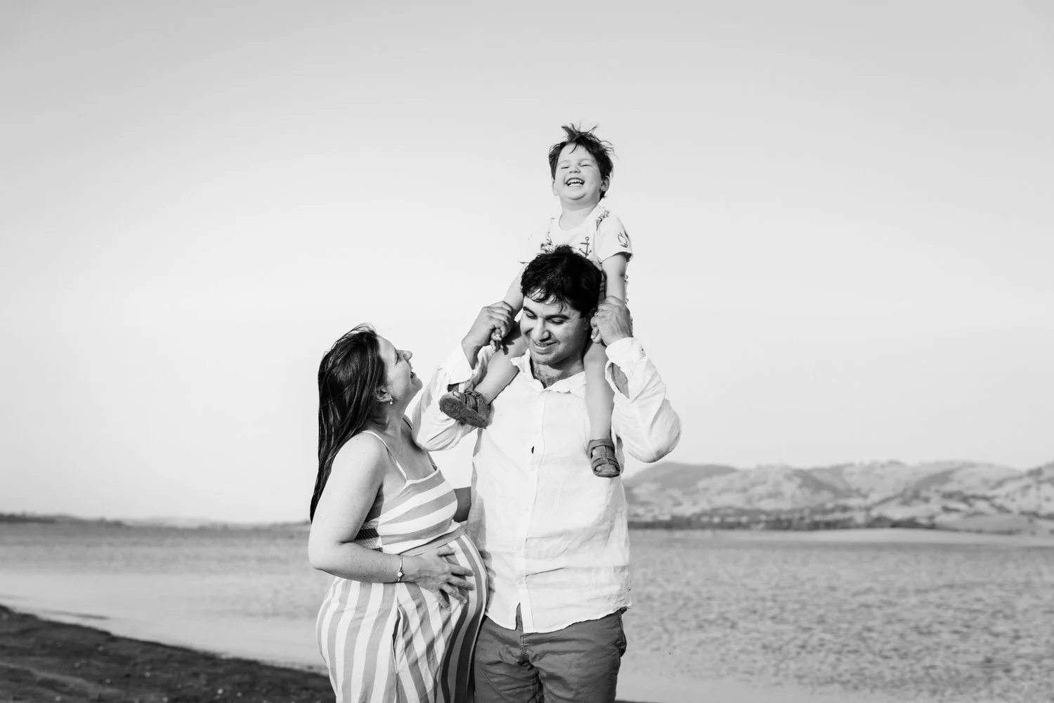 Mum holding her pregnant belly. Toddler laughing on dads shoulders with water behind them