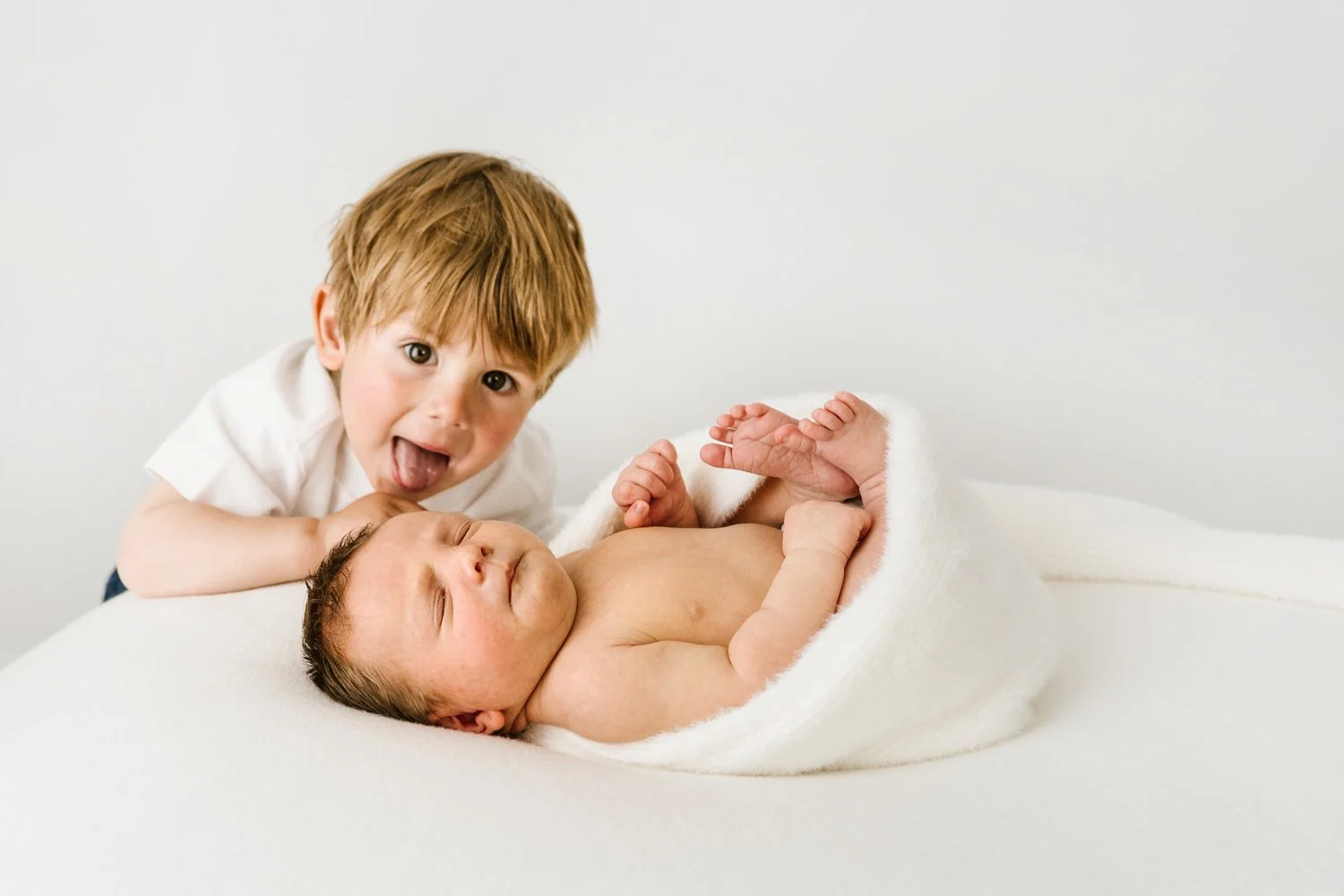 A young boy with light brown hair smiling and sticking his tongue out, lying next to a sleeping newborn wrapped in a white blanket on a white surface.