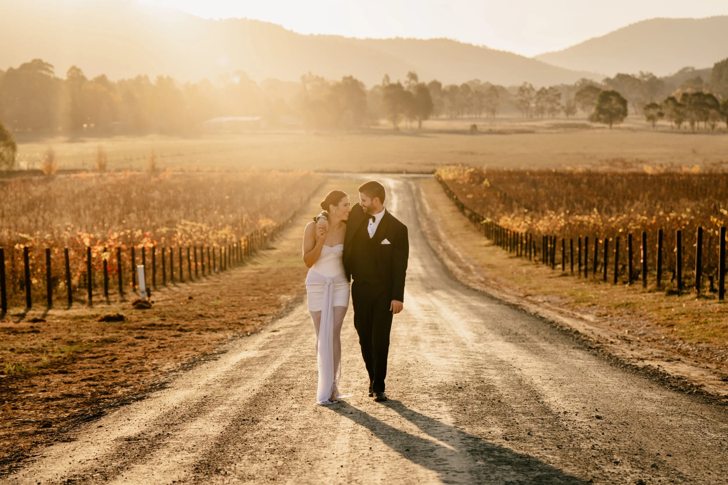 Bride and groom walking down dirt road at sunset