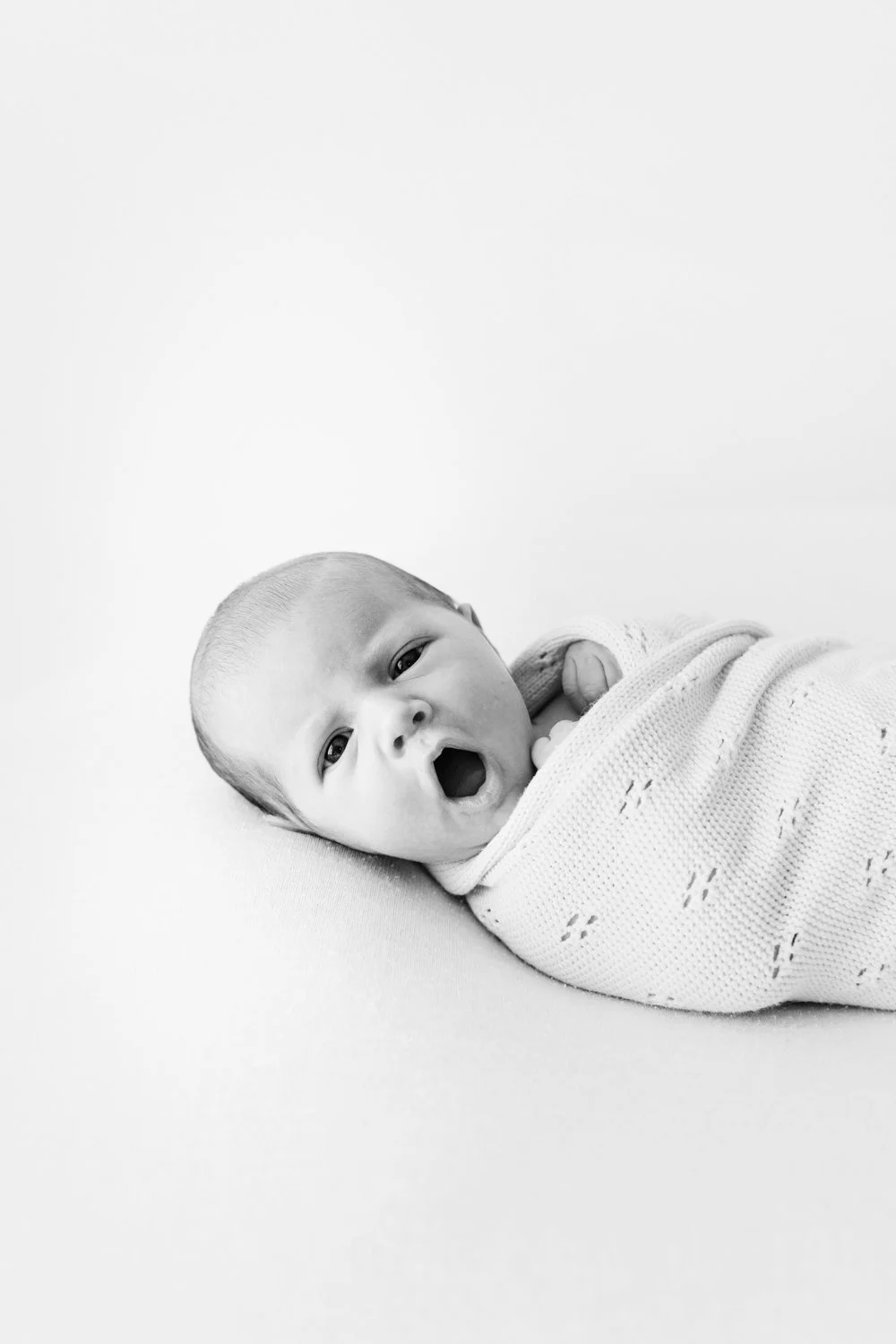 Black and white photo of a yawning baby wrapped in a blanket.