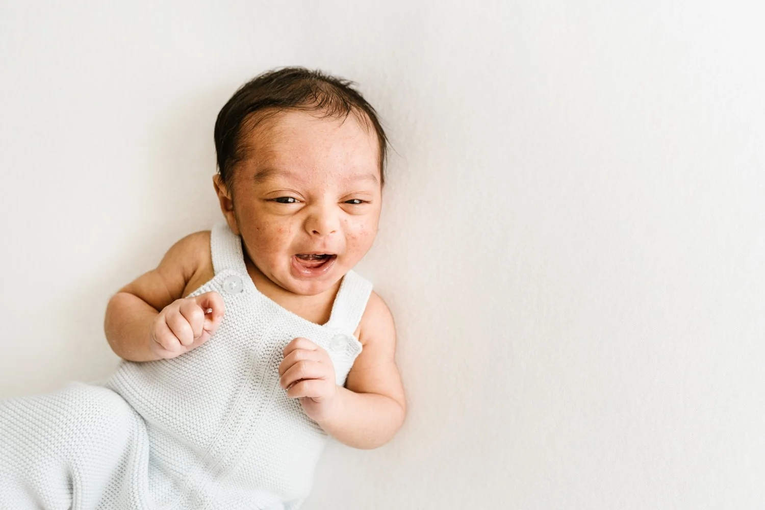 Baby crying or upset, wearing a white sleeveless outfit, lying against a light-colored wall.