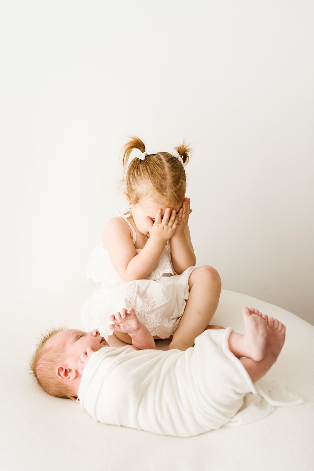 Young girl with pigtails covering her face while sitting on a mattress next to a baby lying on its back.