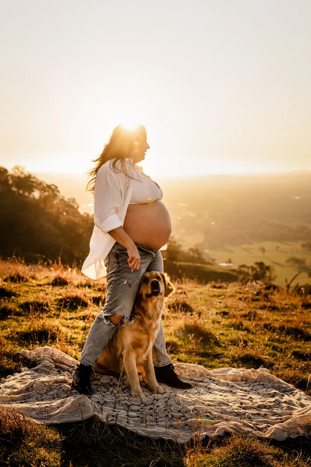 Pregnant woman standing outdoors on a blanket with her dog during sunset.