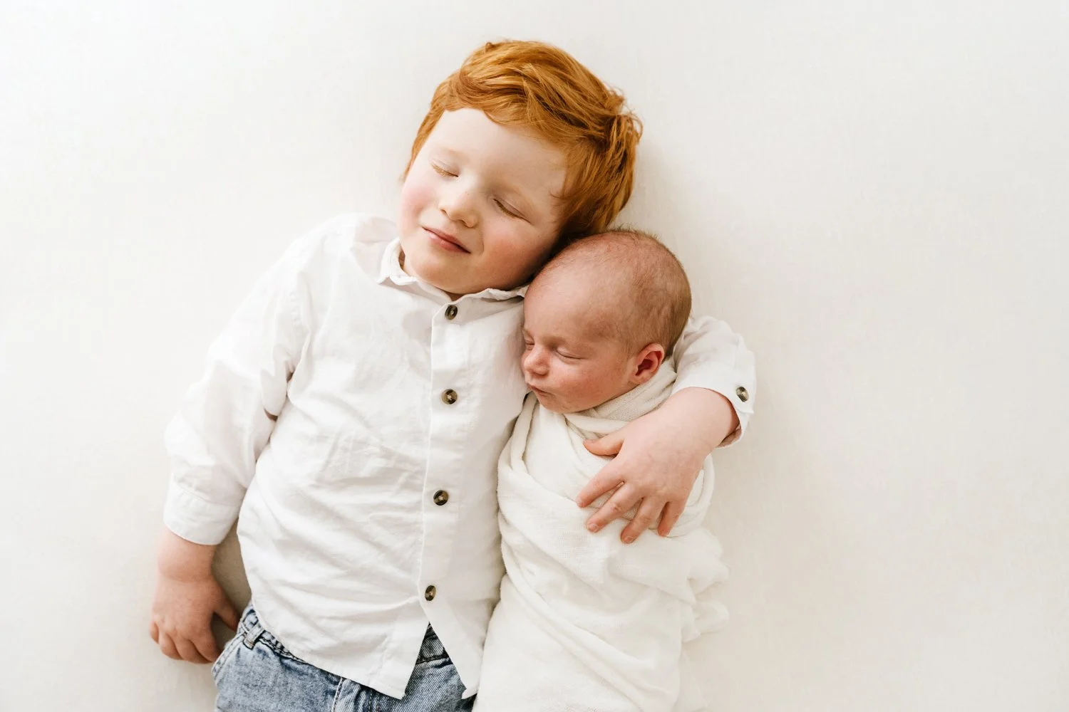 A young red-haired boy hugging a newborn baby wrapped in a white blanket, both with eyes closed, against a plain white background.